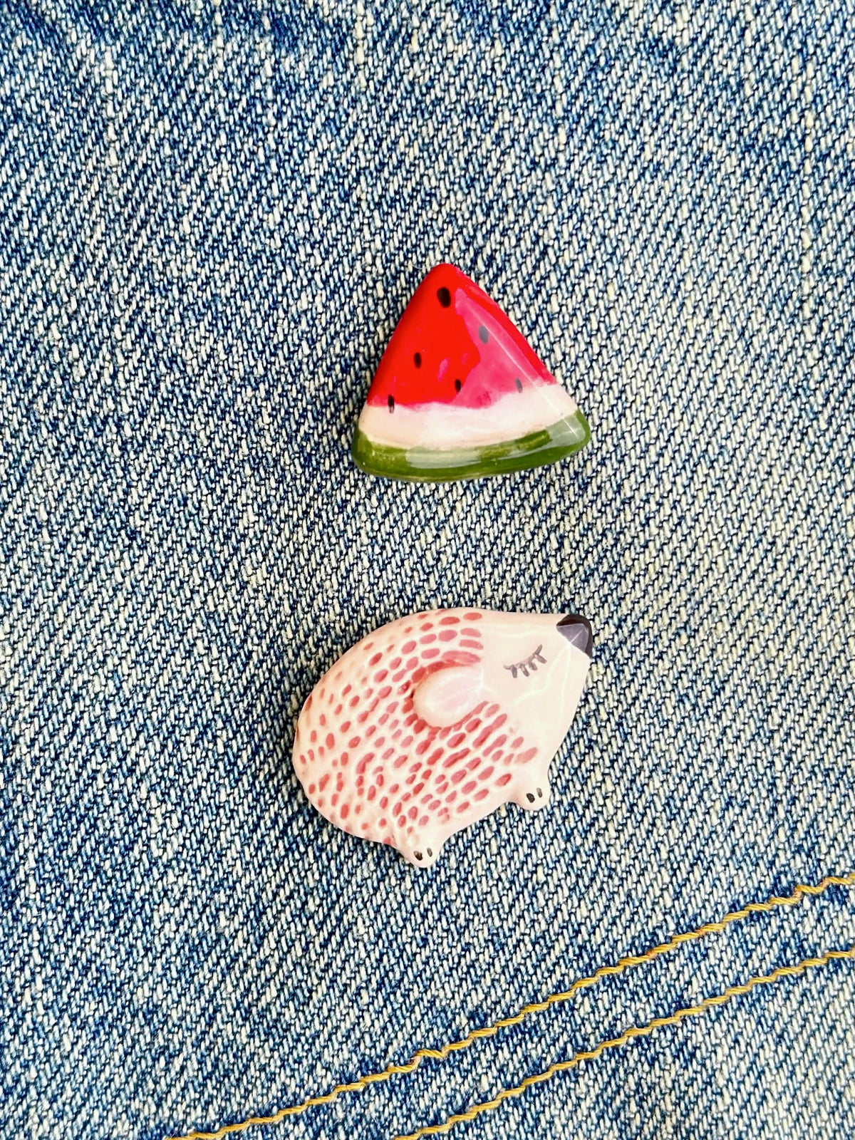 Hand-painted ceramic watermelon slice and sleeping hedgehog brooches pinned to a denim jacket.