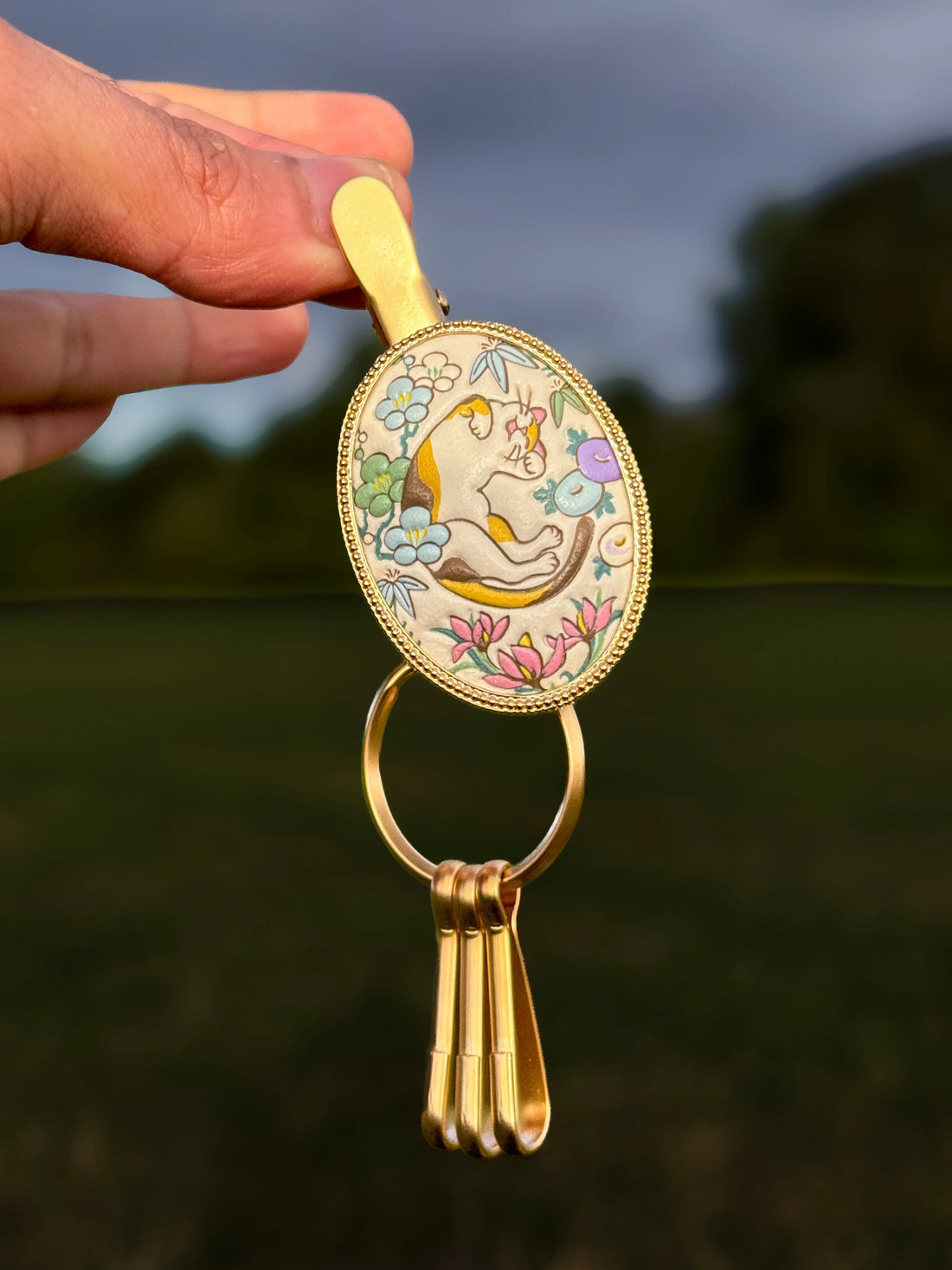 Hand-coloured Yuzen leather key clip with a sleeping calico cat among flowers, held up in a brass frame with three hanging hooks.