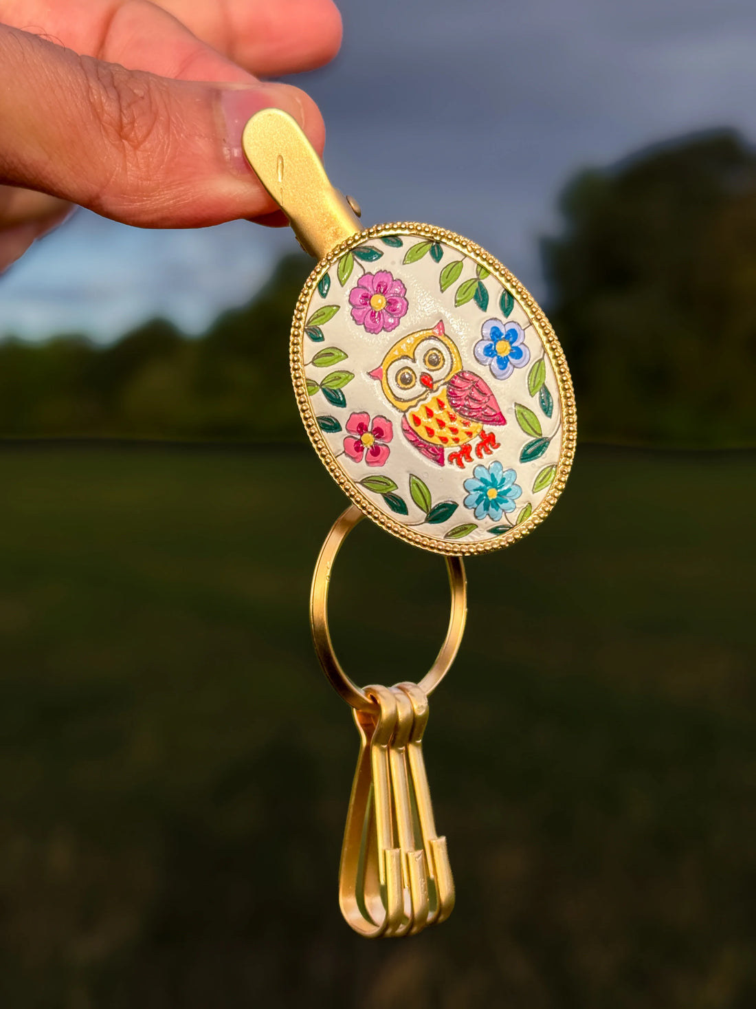 Yuzen leather key clip with a hand-coloured owl among flowers, in a brass frame with three detachable hooks, held outdoors.