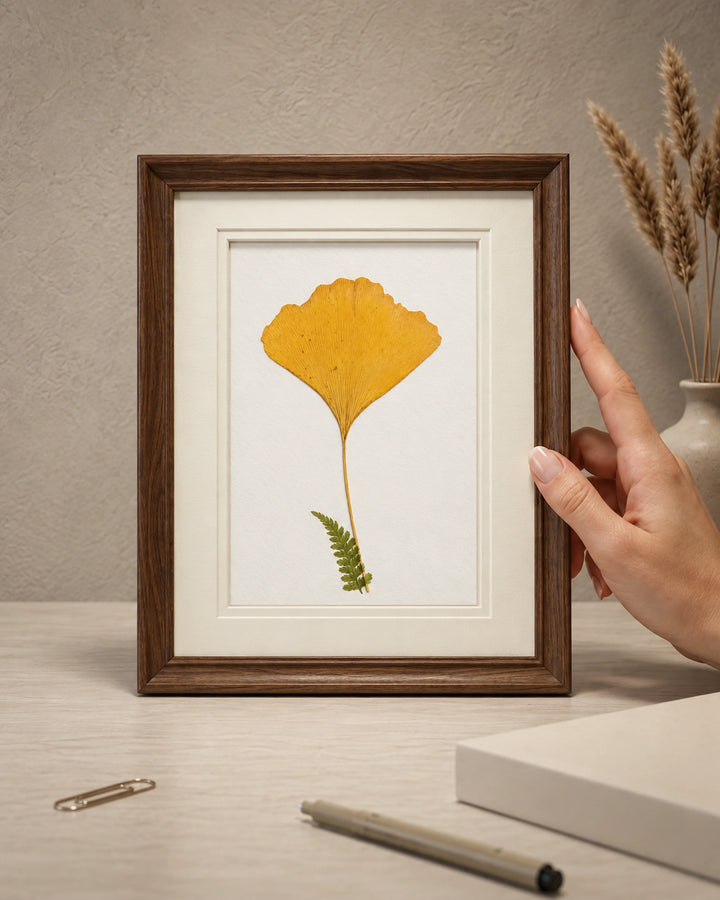 A framed botanical art print featuring a yellow ginkgo leaf and a green fern on a white background, held by a person's hand on a light wood desk with a pen, paperclip, and vase in the background. inside a brown premium frame