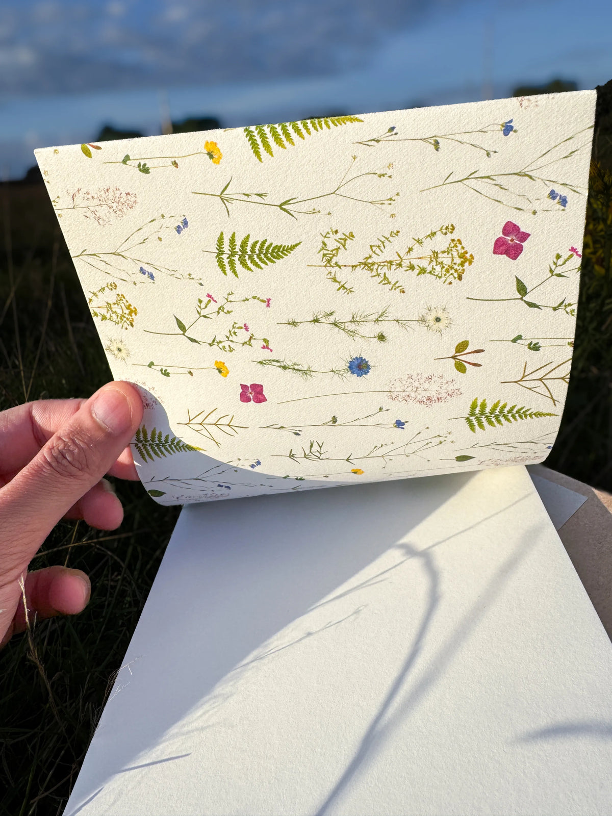 A hand holds a sheet of textured paper printed with a delicate wildflower and fern pattern against an outdoor background of grass and sky.