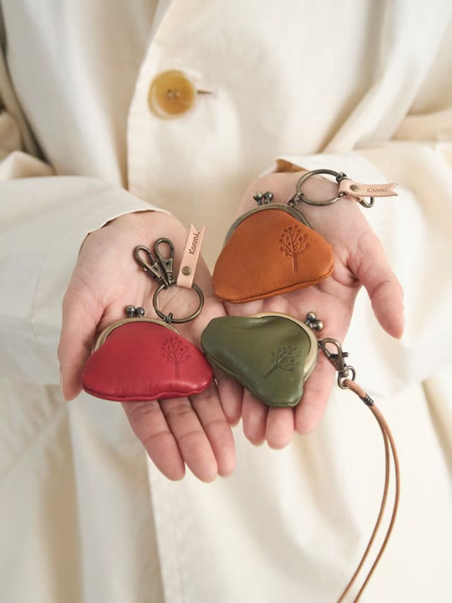 Three small leather coin purses in red, orange-brown, and olive green are held in two hands against a cream-colored coat. Each purse has a metal clasp closure, a key ring, and a small leather tag.