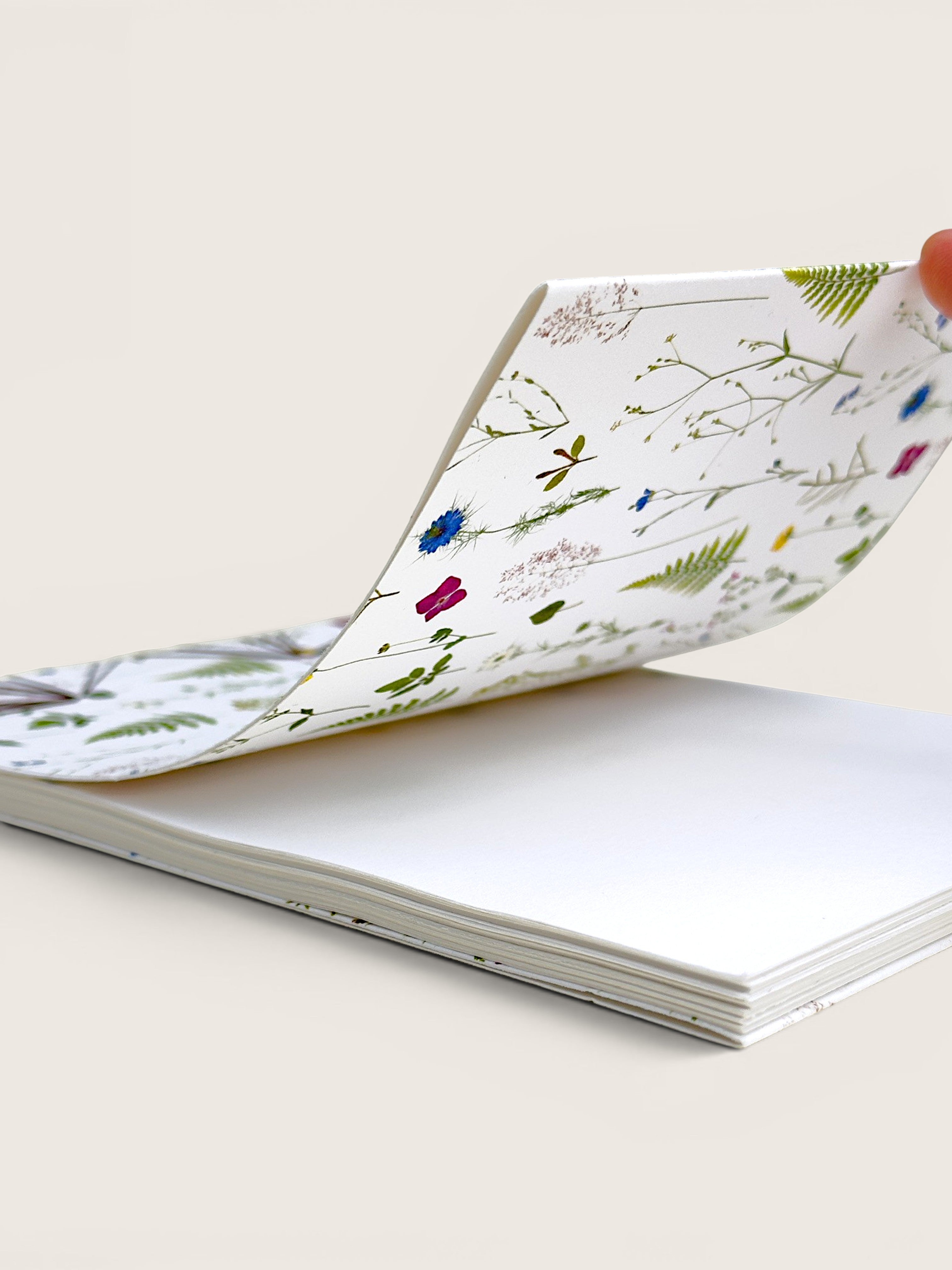 A hardcover notebook with a white cover decorated with delicate botanical illustrations, shown open with a hand lifting one page to reveal blank white interior pages. hand made in south west france