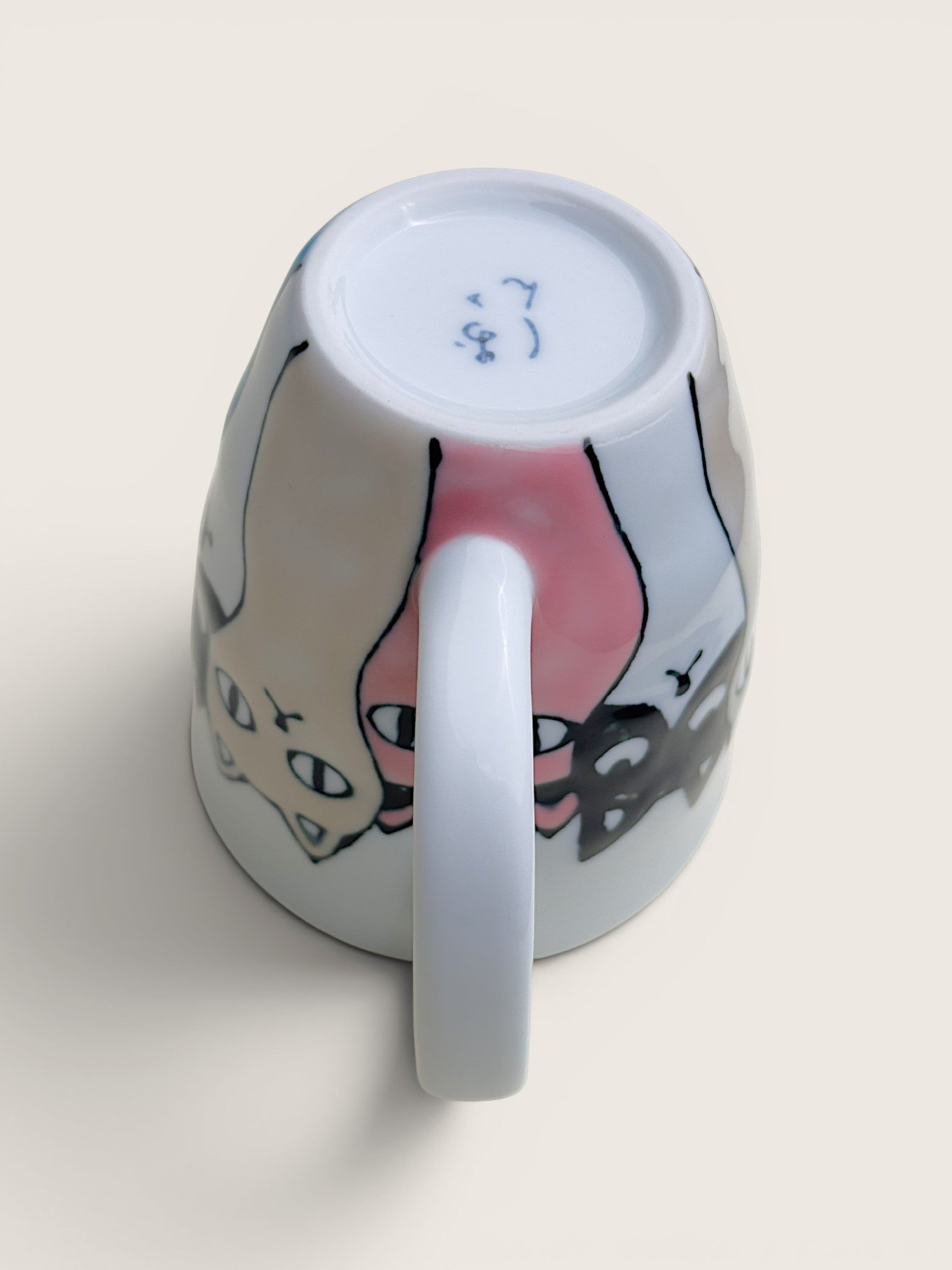 A white ceramic mug shown from an overhead angle, revealing a hand-drawn style illustration of two cats on the outside and blue maker's marks on the bottom of the cup.