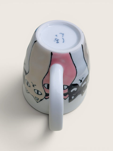 A white ceramic mug shown from an overhead angle, revealing a hand-drawn style illustration of two cats on the outside and blue maker's marks on the bottom of the cup.