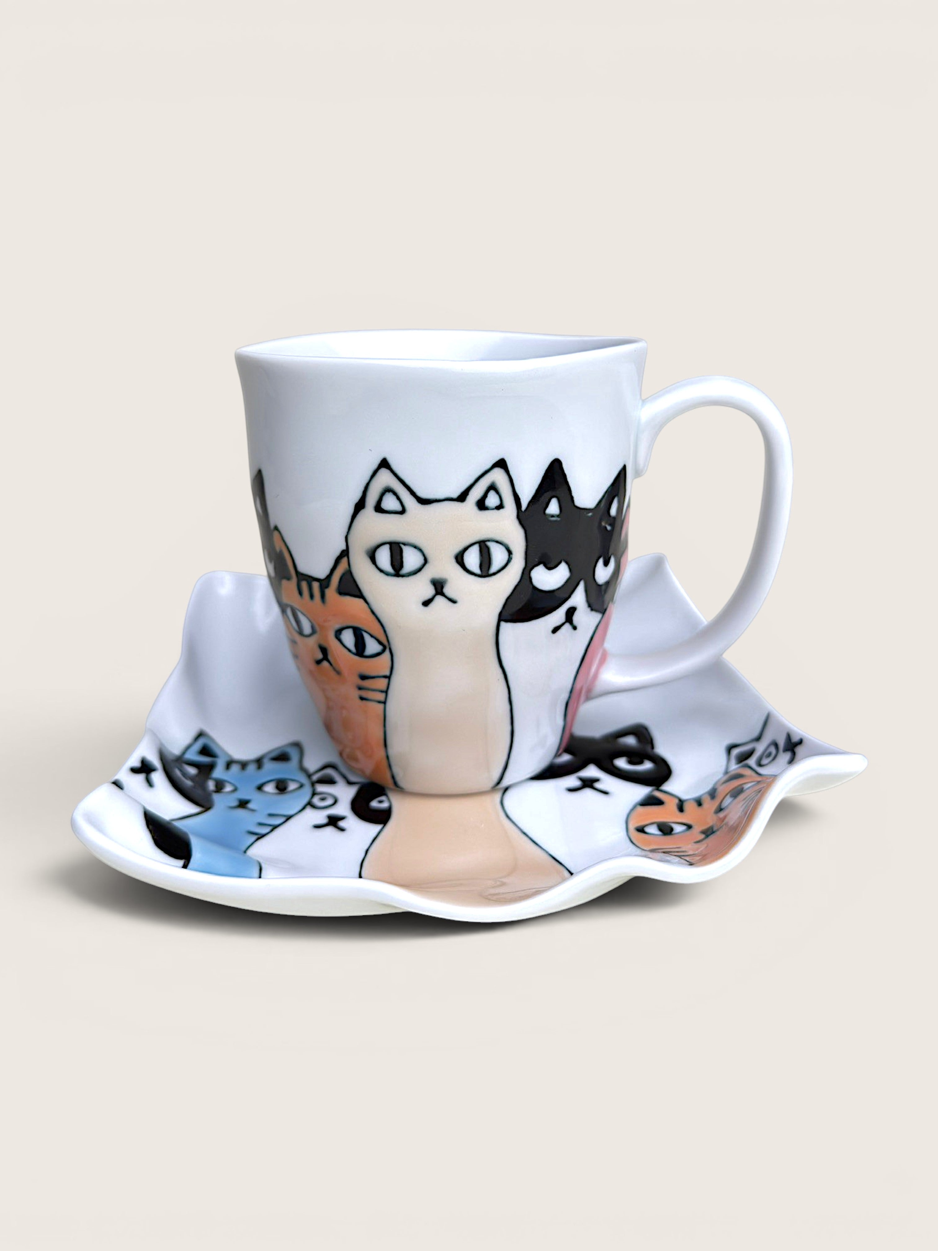 A white ceramic coffee mug and matching saucer set, both decorated with a whimsical illustration of various cat faces in different colors including beige, black, orange, and blue.