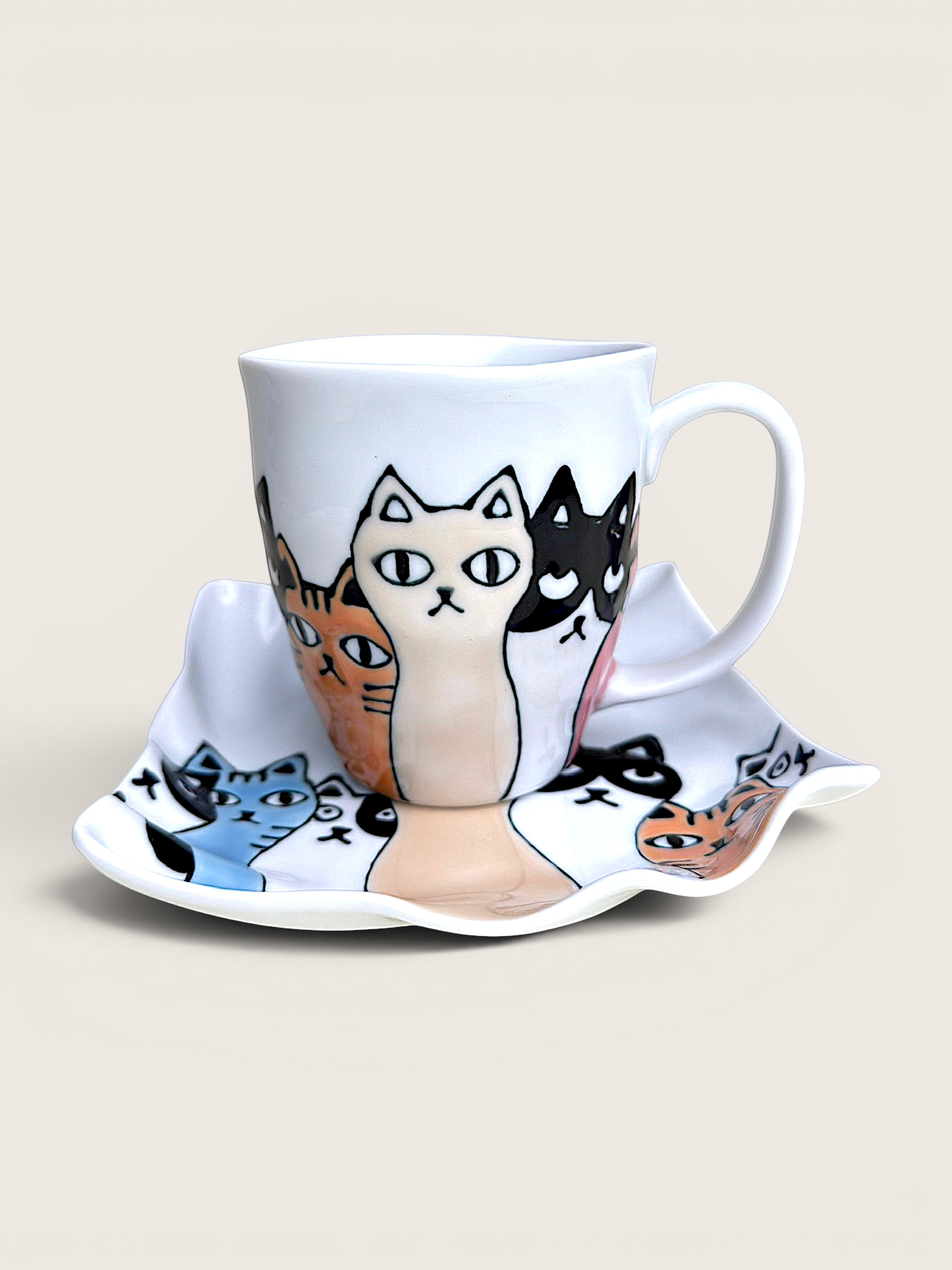 A white ceramic coffee mug and matching saucer set, both decorated with a whimsical illustration of multiple cartoon cats in various colors like black, orange, blue, and cream, set against a solid light beige background.