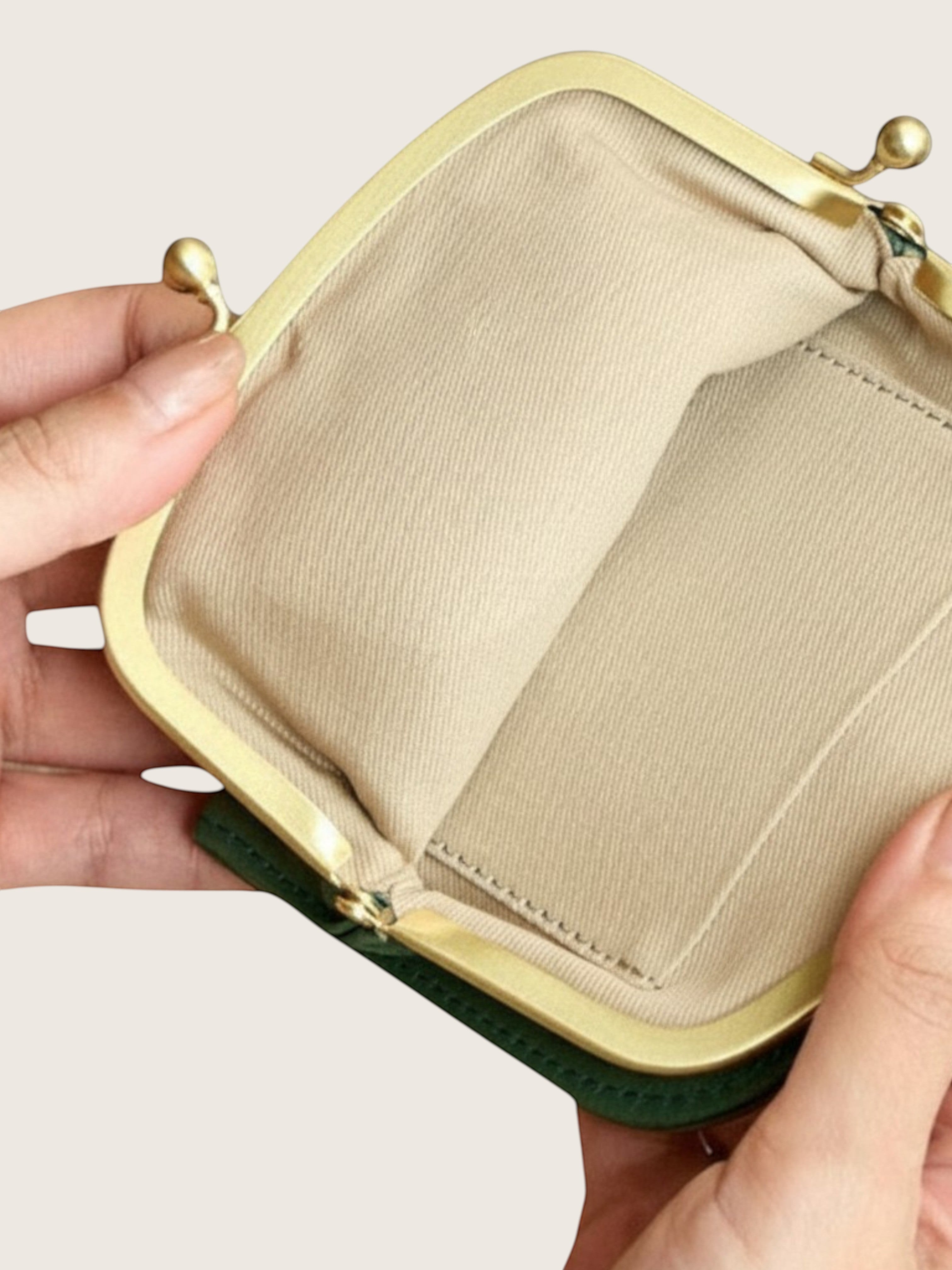 An open coin purse or small clutch wallet held by two hands, showing a beige fabric interior, gold-tone metal frame, and a dark green exterior edge against a light neutral background.