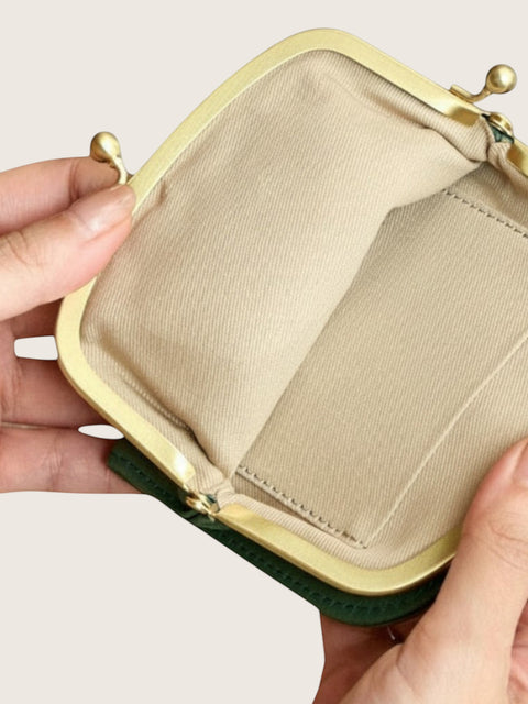 An open coin purse or small clutch wallet held by two hands, showing a beige fabric interior, gold-tone metal frame, and a dark green exterior edge against a light neutral background.