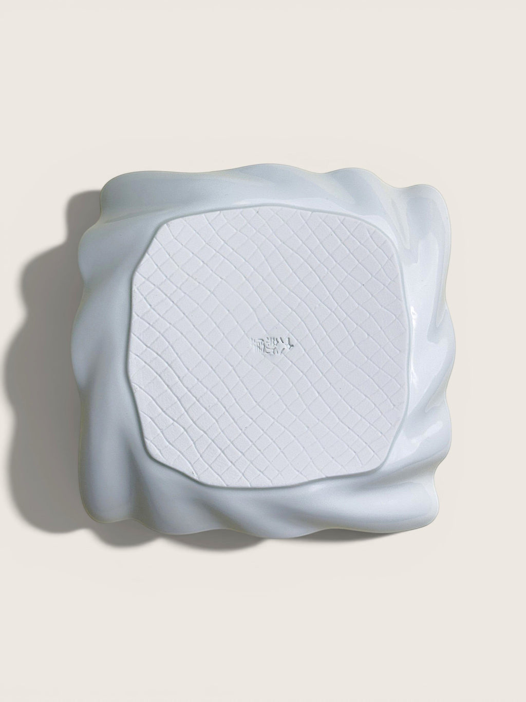 A white textured square mat with a wavy, sculptural raised border, photographed from directly above on a light beige background.