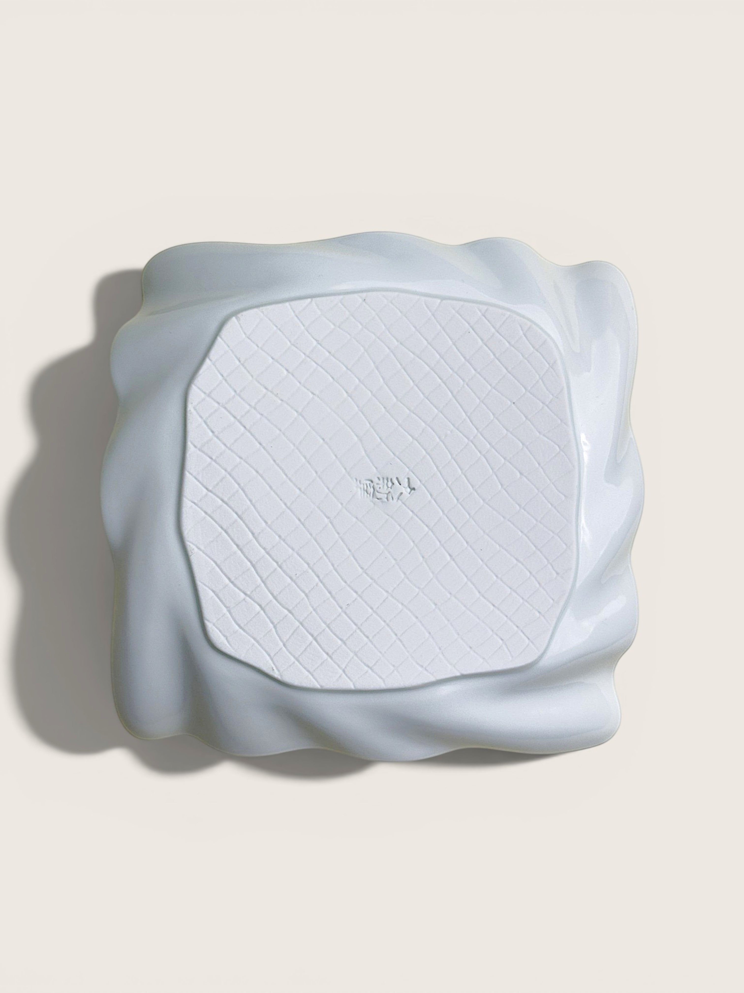 A white textured square mat with a wavy, sculptural raised border, photographed from directly above on a light beige background.