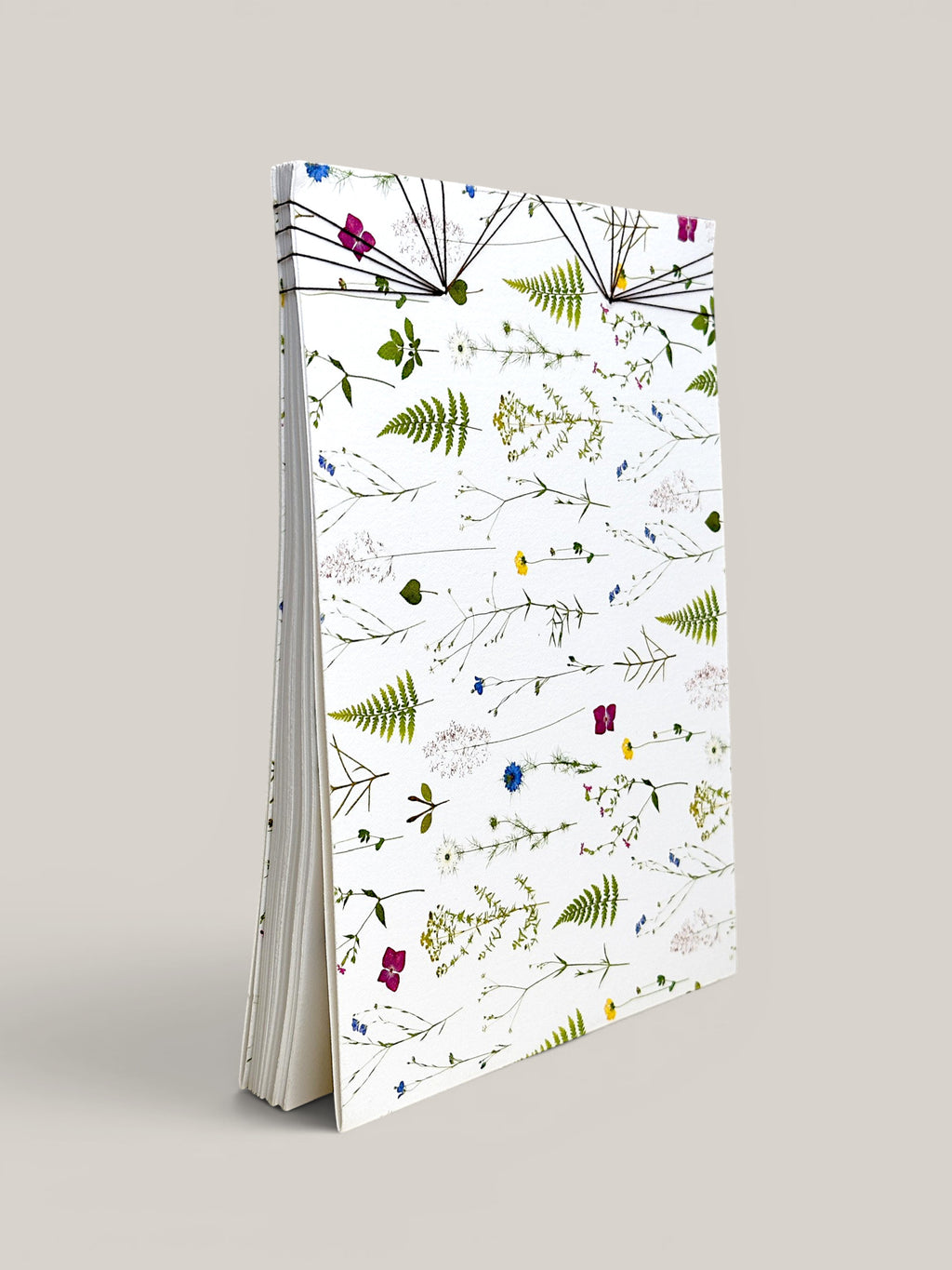 A tall, slim notebook or journal standing upright on a light gray background. The cover is white with an all-over botanical pattern of green ferns and small multicolored wildflowers, and the pages are visible along the left side. hand made in south west france