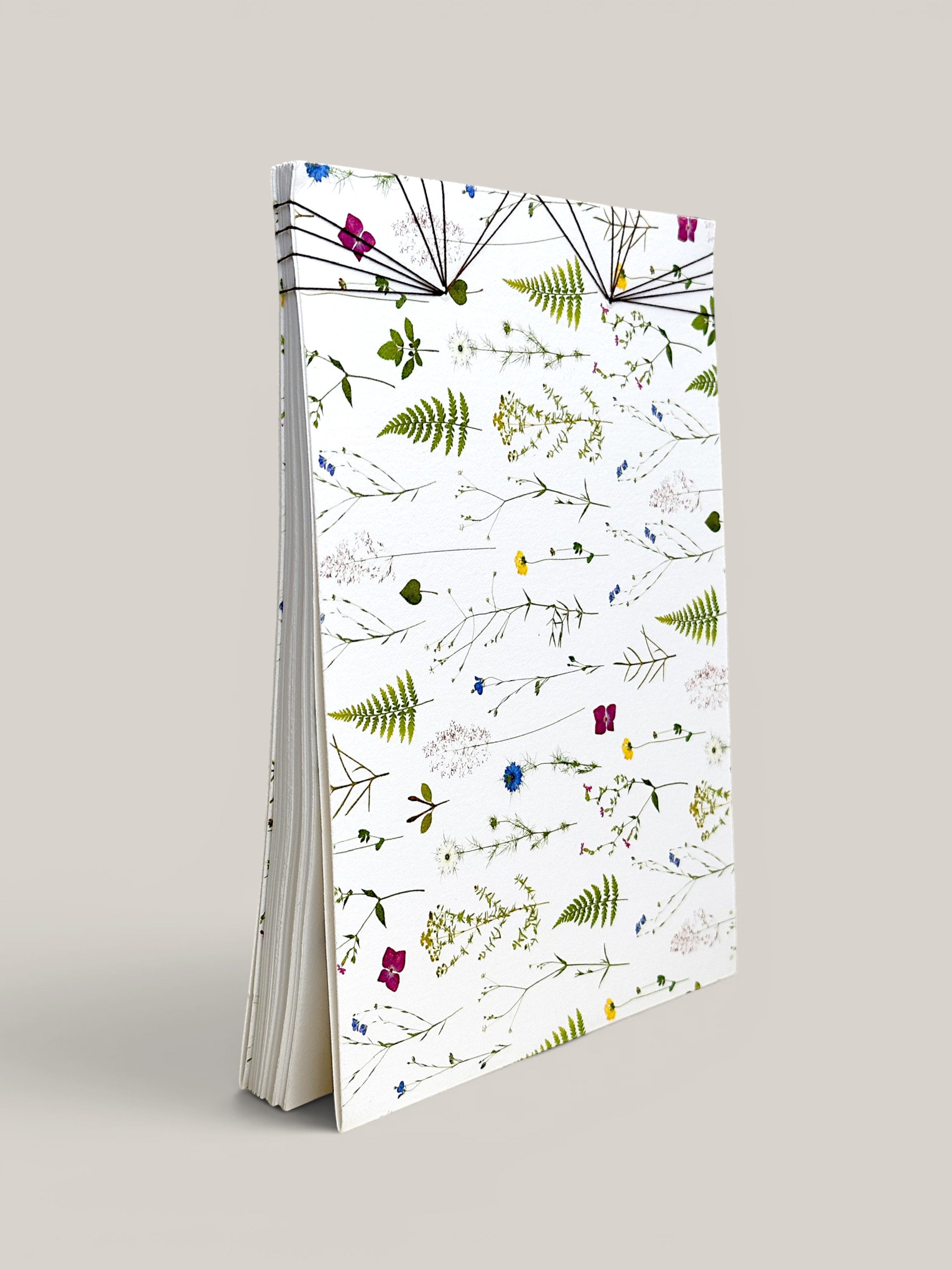 A tall, slim notebook or journal standing upright on a light gray background. The cover is white with an all-over botanical pattern of green ferns and small multicolored wildflowers, and the pages are visible along the left side. hand made in south west france