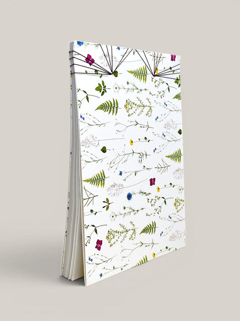 A tall, slim notebook or journal standing upright on a light gray background. The cover is white with an all-over botanical pattern of green ferns and small multicolored wildflowers, and the pages are visible along the left side. hand made in south west france