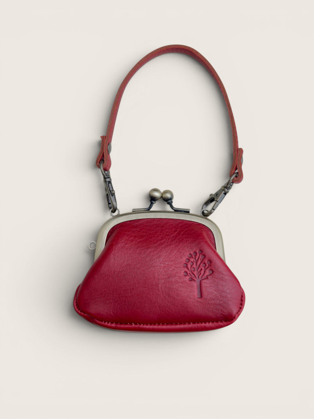 A small red leather kiss-lock coin purse with a matching red shoulder strap, silver-tone metal frame, and an embossed tree logo on the front, shown against a light beige background.