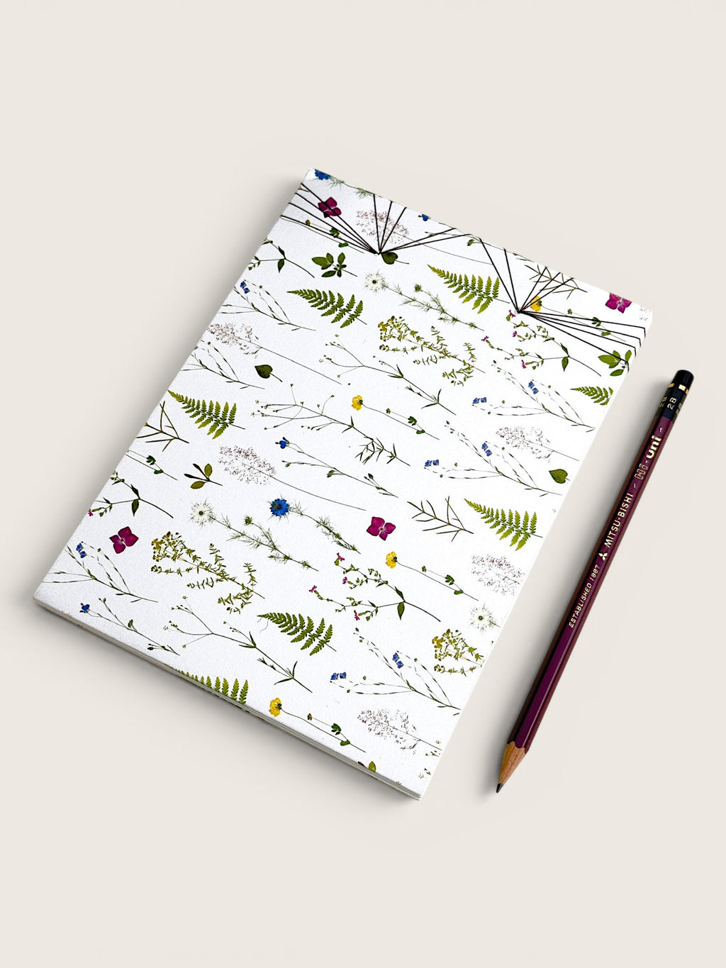 A notebook with a white cover decorated with delicate botanical illustrations, shown next to a dark purple pencil on a light beige background. hand made in south west france