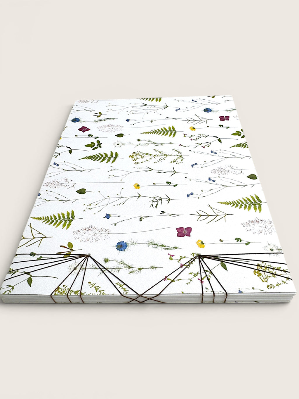 A stack of white paper notebooks or cards featuring a delicate botanical pattern of pressed flowers and leaves, bound together with black string at the bottom, set against a solid light beige background.