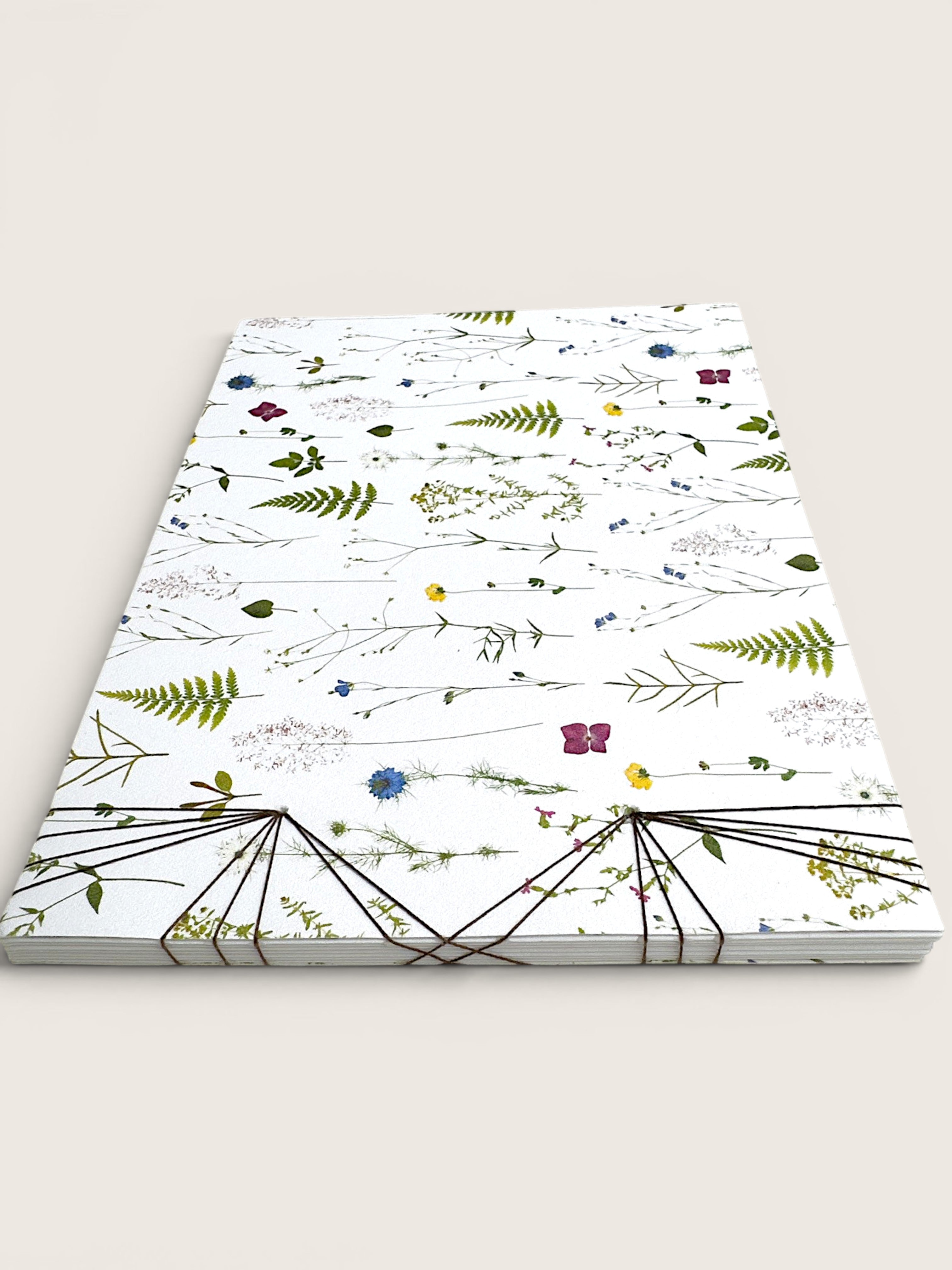 A stack of white paper notebooks or cards featuring a delicate botanical pattern of pressed flowers and leaves, bound together with black string at the bottom, set against a solid light beige background.
