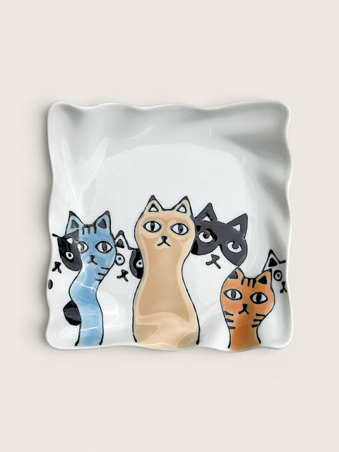 A square white ceramic dish with wavy edges, decorated with a row of stylized cartoon cat heads in blue, beige, black, and orange, set against a solid light beige background.