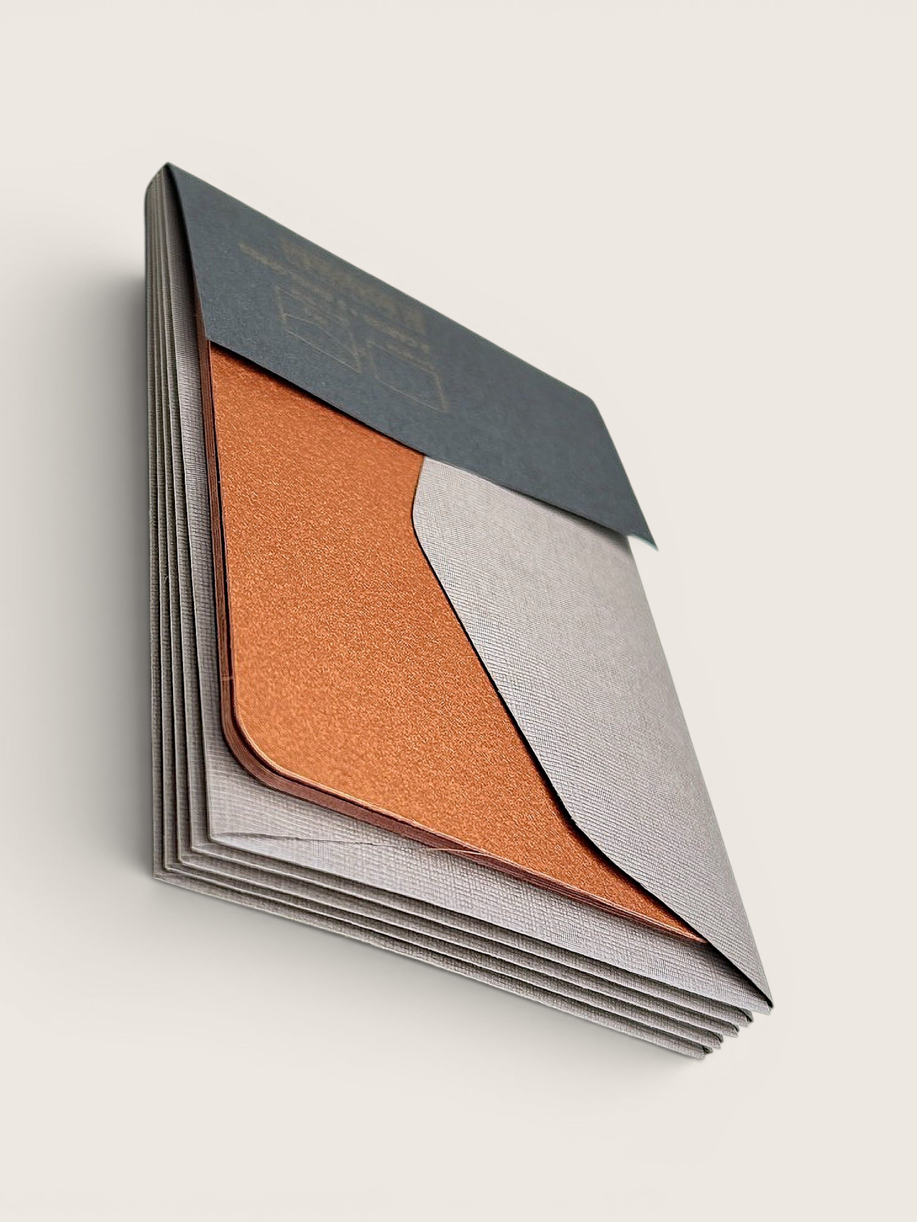 A stack of textured paper envelopes or cardstock sheets with overlapping panels in dark charcoal, metallic copper, and light gray, photographed on a clean off-white background.