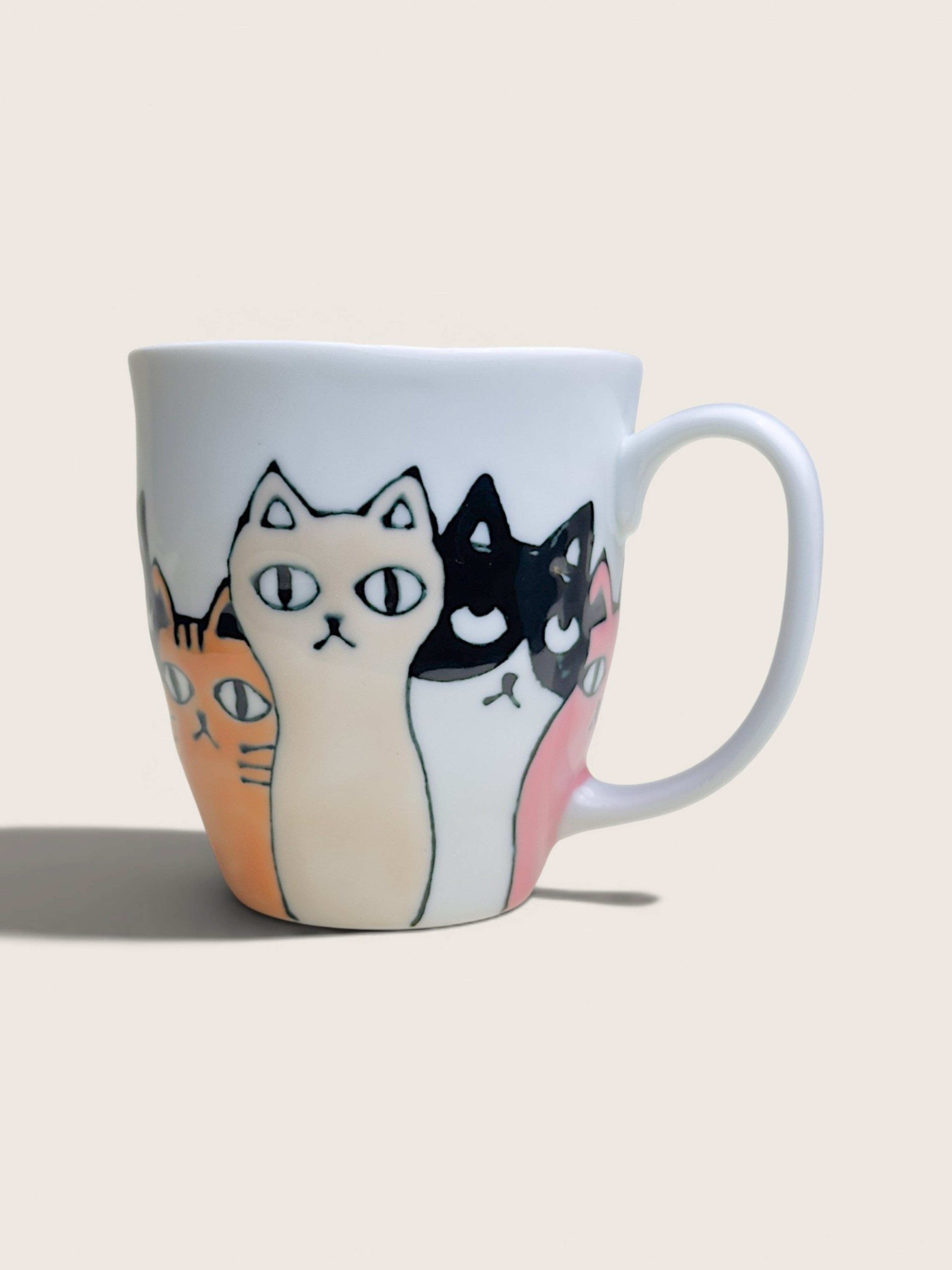 A white ceramic coffee mug featuring a whimsical illustration of several cats in different colors (cream, black, orange, pink) peeking out from behind the front cat, set against a solid light beige background.