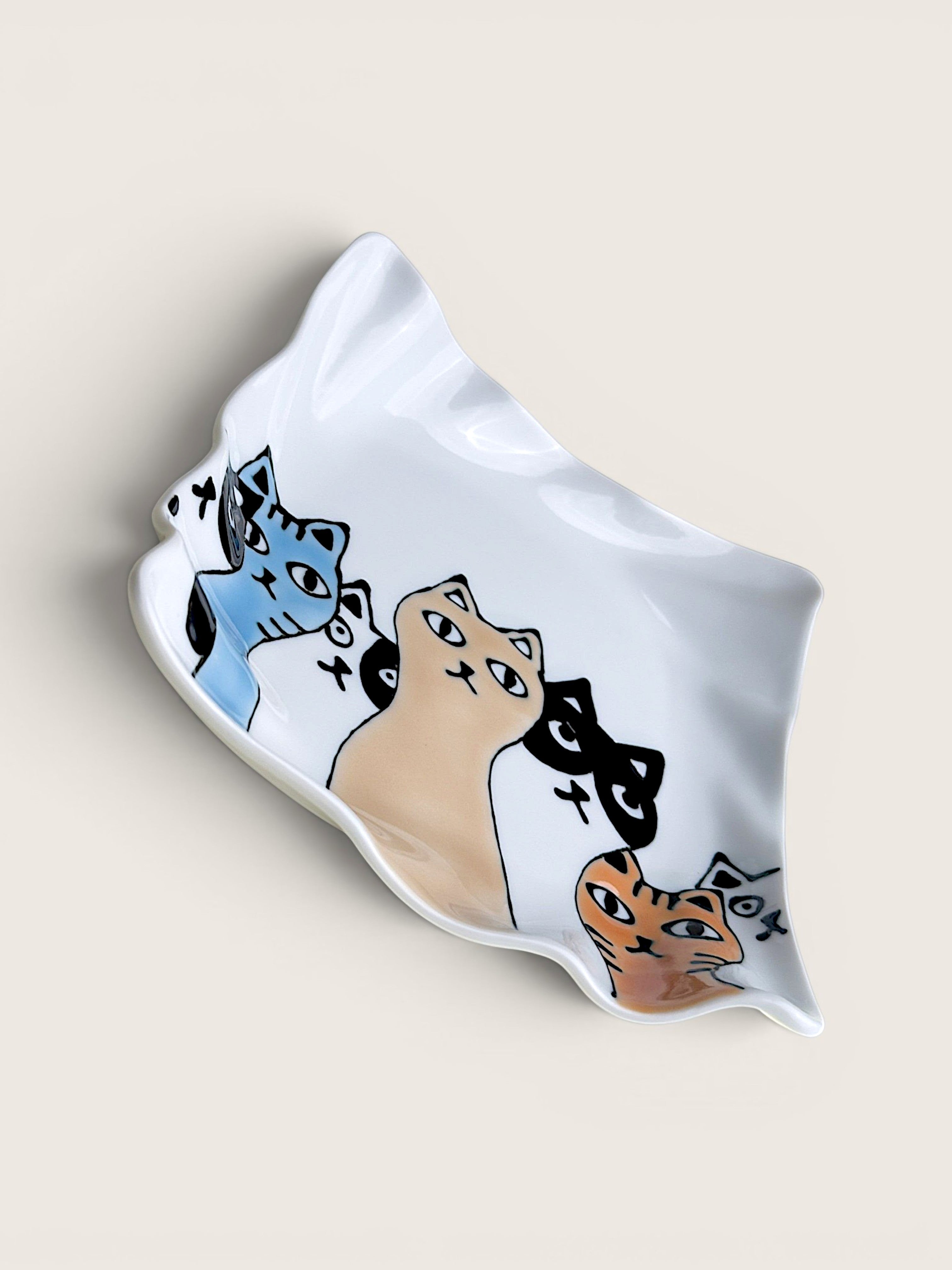 A white ceramic dish with an organic, wavy shape, decorated with a hand-drawn style illustration of four cats in blue, beige, black, and orange. The dish is photographed on a solid, warm off-white background.