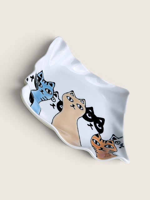 A white ceramic dish with an organic, wavy shape, decorated with a hand-drawn style illustration of four cats in blue, beige, black, and orange. The dish is photographed on a solid, warm off-white background.