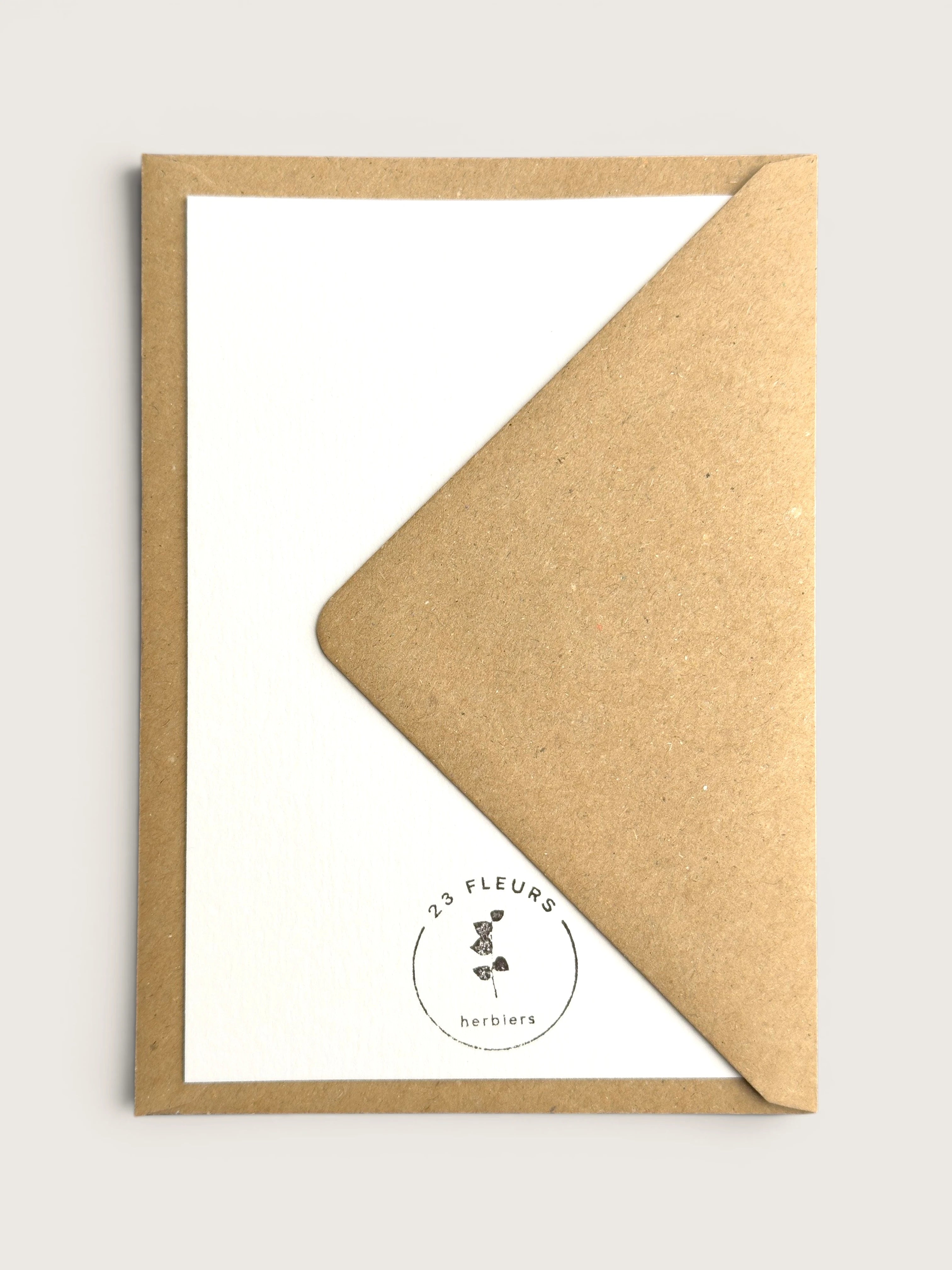 A kraft brown envelope partially covering a white card with a small circular '23 Fleurs herbiers' logo printed near the bottom.