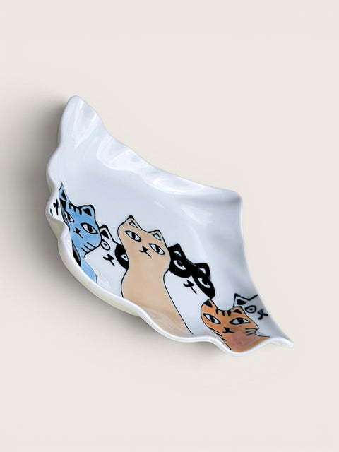 A white ceramic dish with a wavy, shell-like edge, decorated with hand-painted cartoon cats in blue, beige, black, and orange, set against a solid light beige background.
