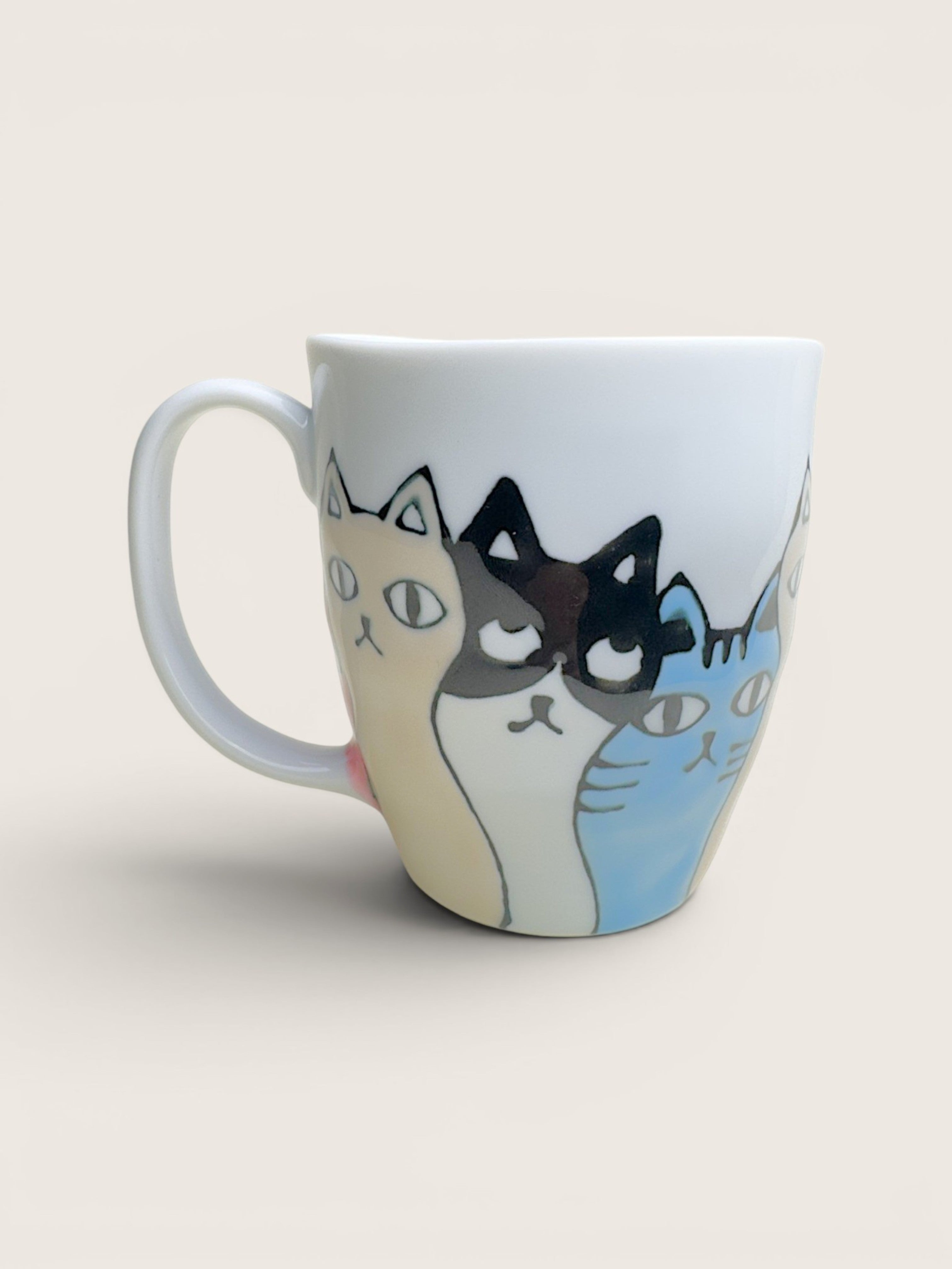 A white ceramic coffee mug featuring a whimsical illustration of three cats with different colorways (cream, black-and-white, and blue) peeking out from behind the front cat, set against a solid light beige background.