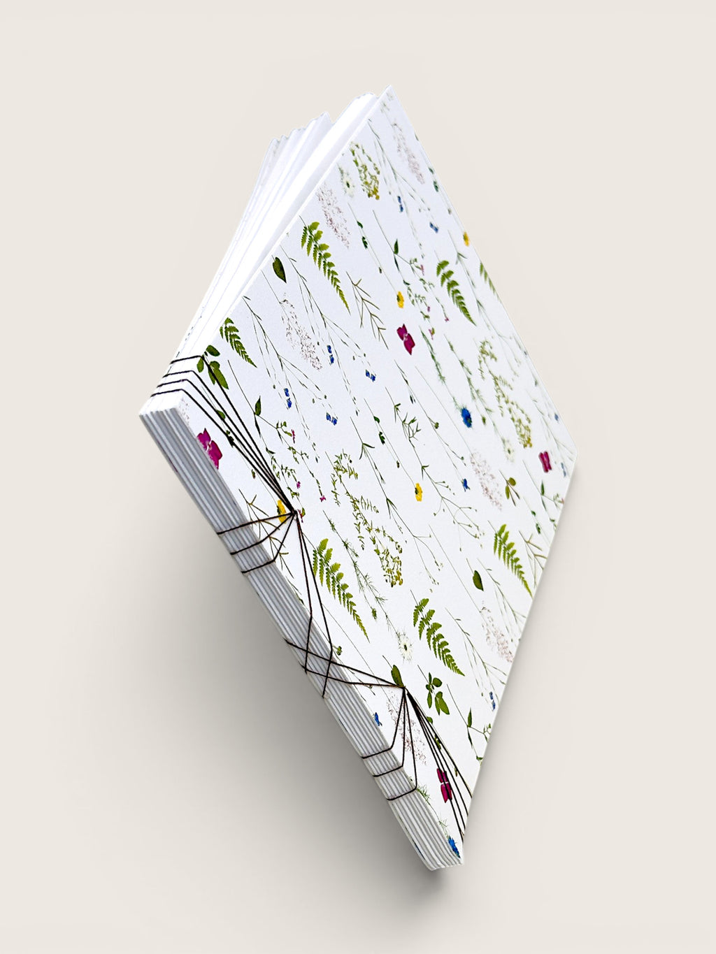 A stack of patterned paper napkins or small paper sheets bound with black string, shown at an angle on a light beige background. The sheets have a white base with scattered botanical illustrations in green, blue, yellow, and pink.