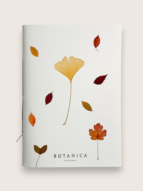 A Botanica L'Automne notebook with a white cover decorated with watercolor autumn leaves in shades of orange, yellow, and red, set against a light beige background.