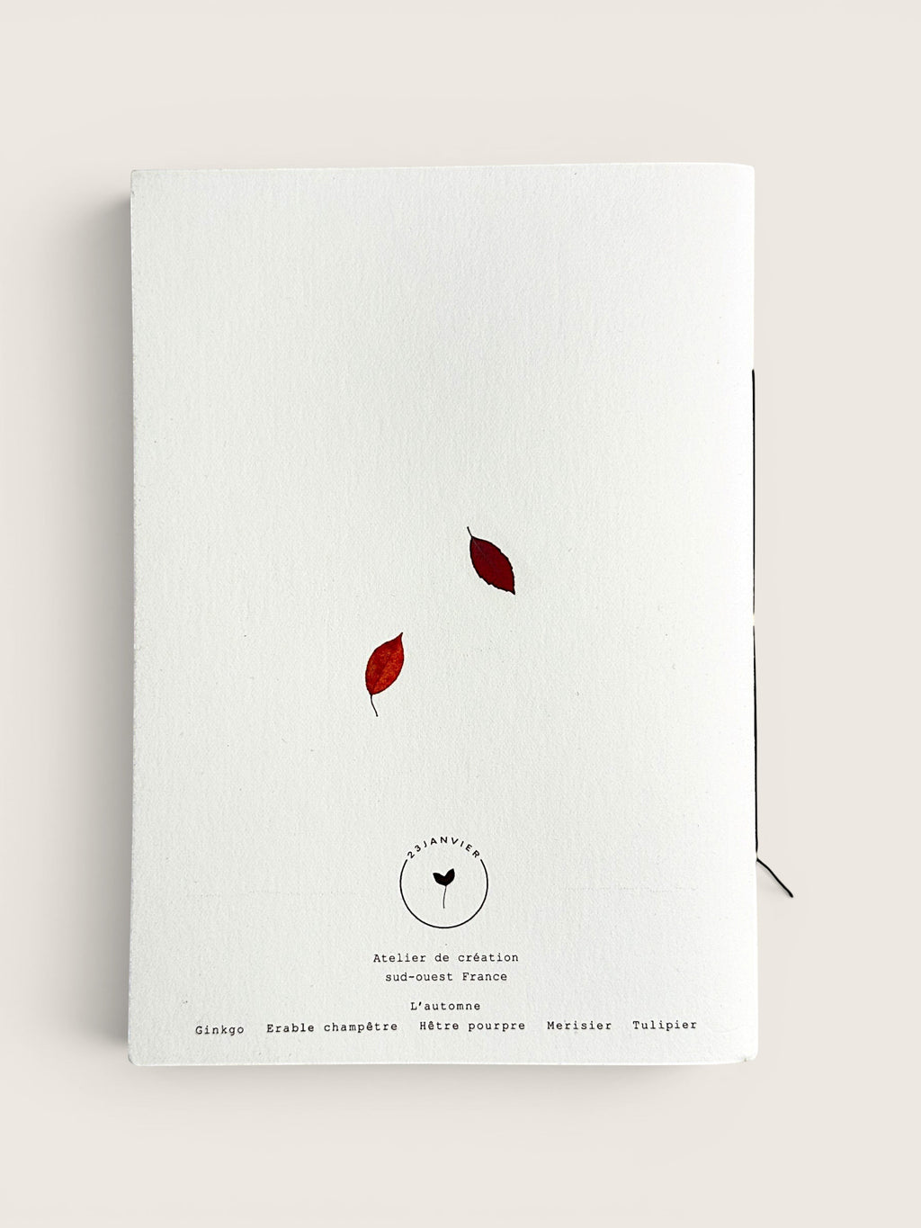 A white textured paper notebook or journal with two small red leaves and black printed text on the cover, photographed against a light beige background.