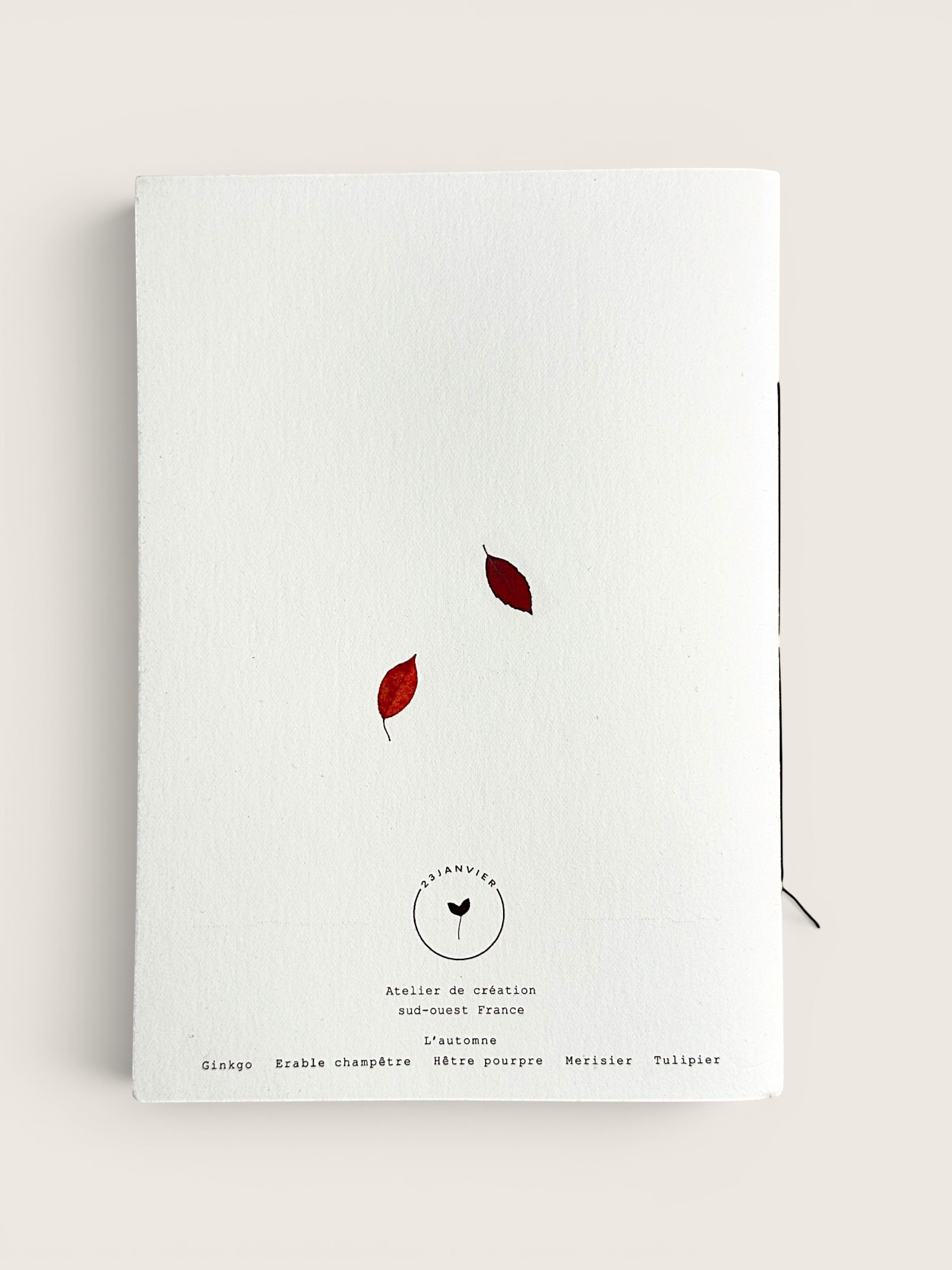 A white textured paper notebook or journal with two small red leaves and black printed text on the cover, photographed against a light beige background.