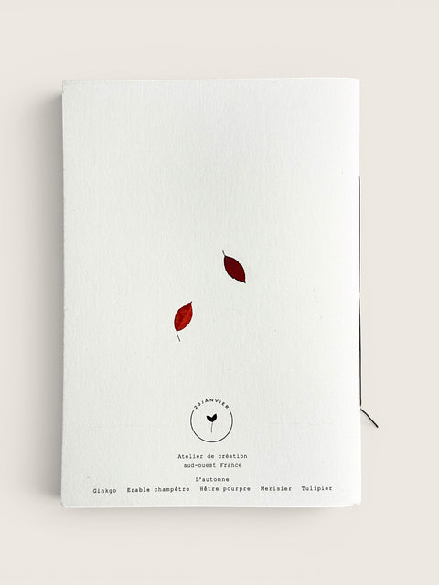 A white textured paper notebook or journal with two small red leaves and black printed text on the cover, photographed against a light beige background.