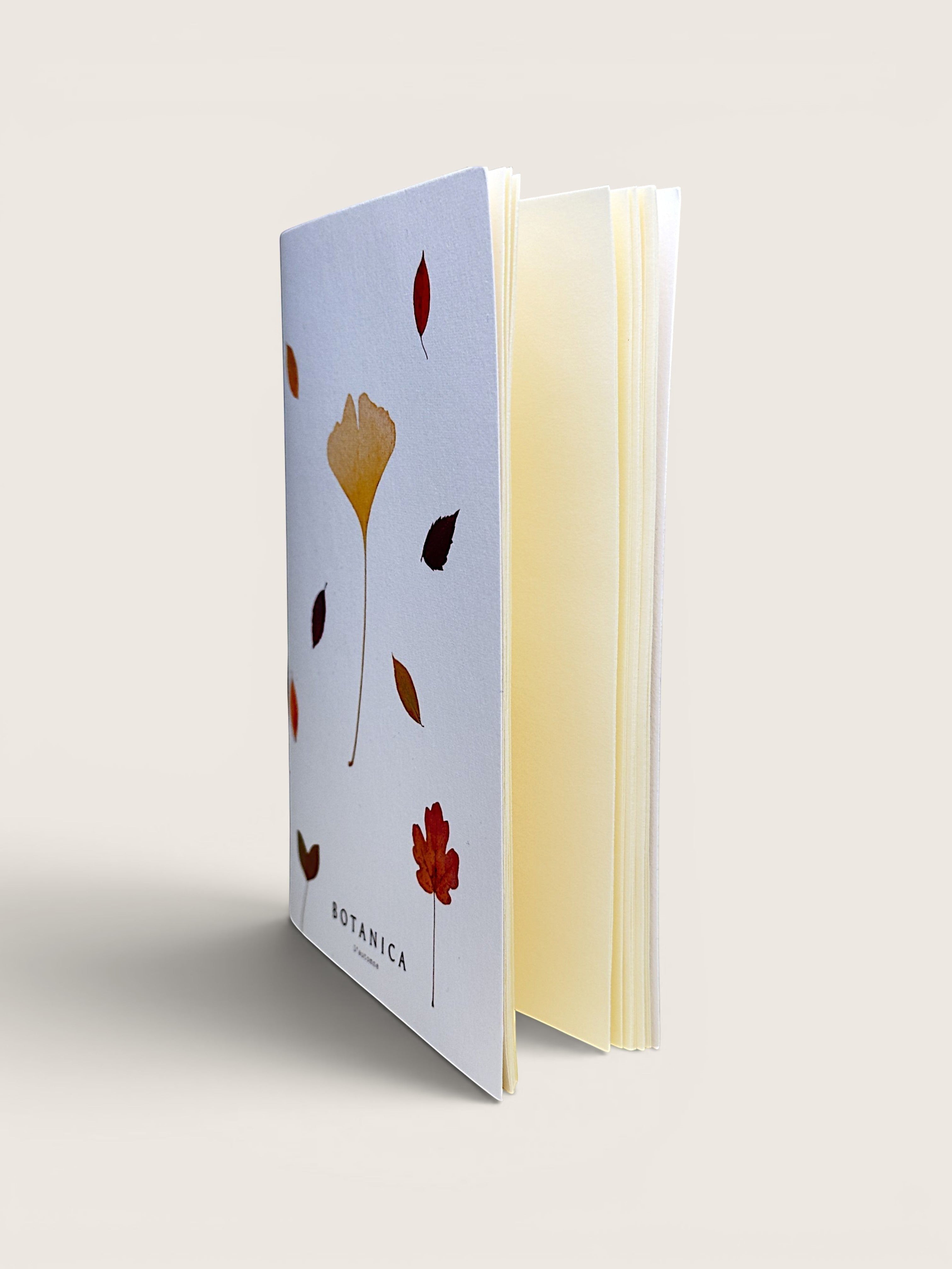 A botanical art journal standing upright at an angle, showing a white cover decorated with gold, red, and brown leaf illustrations, and cream-colored pages visible on the right side, set against a solid light beige background.
