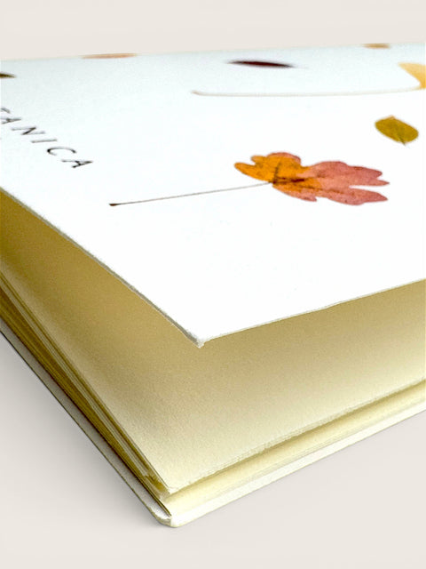 A close-up of a cream-colored notebook or journal with a white cover decorated with a pressed autumn leaf and the partial brand name 'LANICA'.