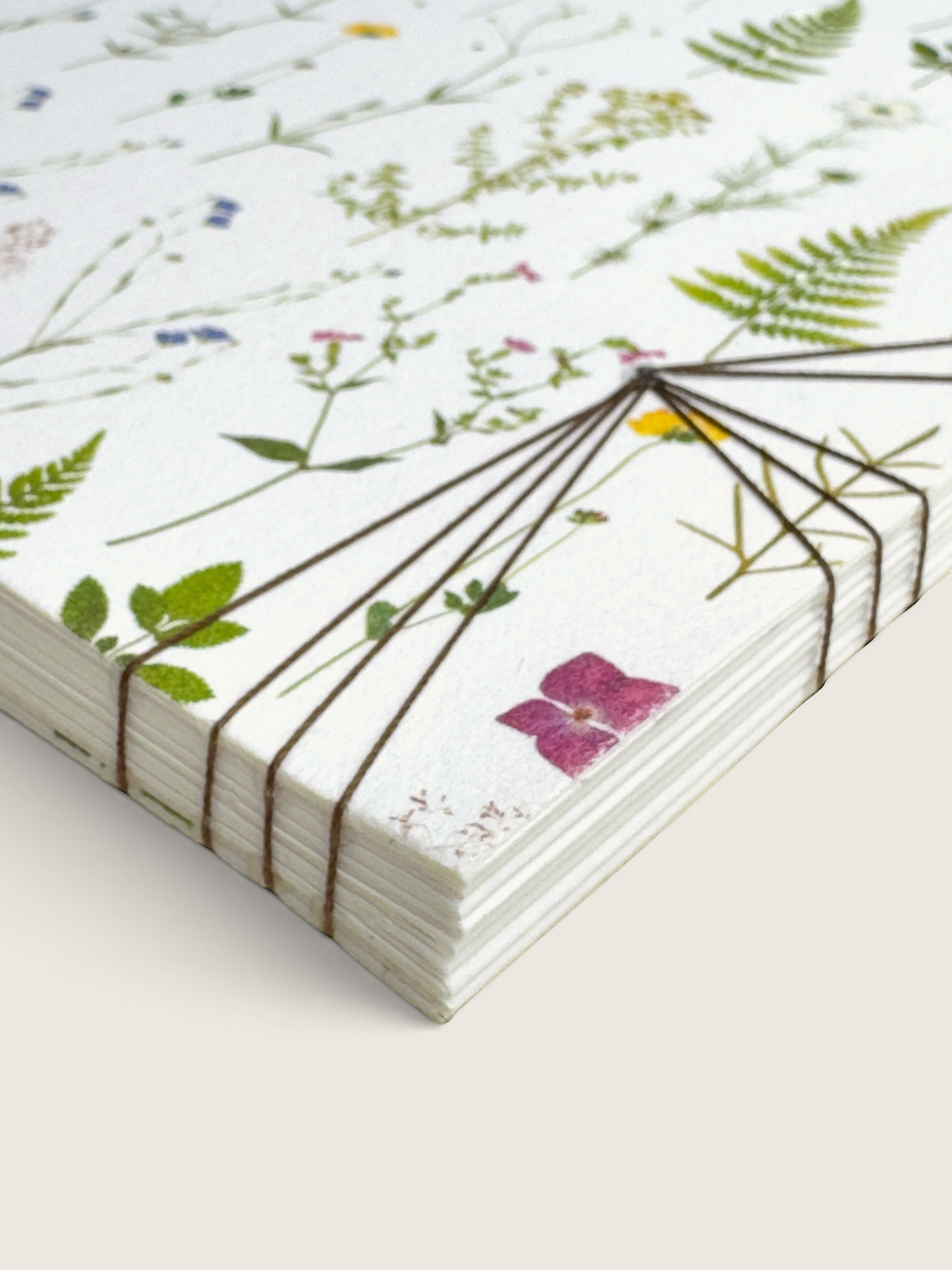 A stack of white paper sheets with a delicate botanical and wildflower print, bound together with thin dark elastic cords, shown on a light beige background.