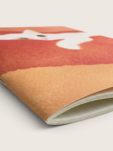 A close-up of a notebook or journal with a textured cover featuring a stylized white fox on a terracotta and tan background, photographed on a light neutral surface.