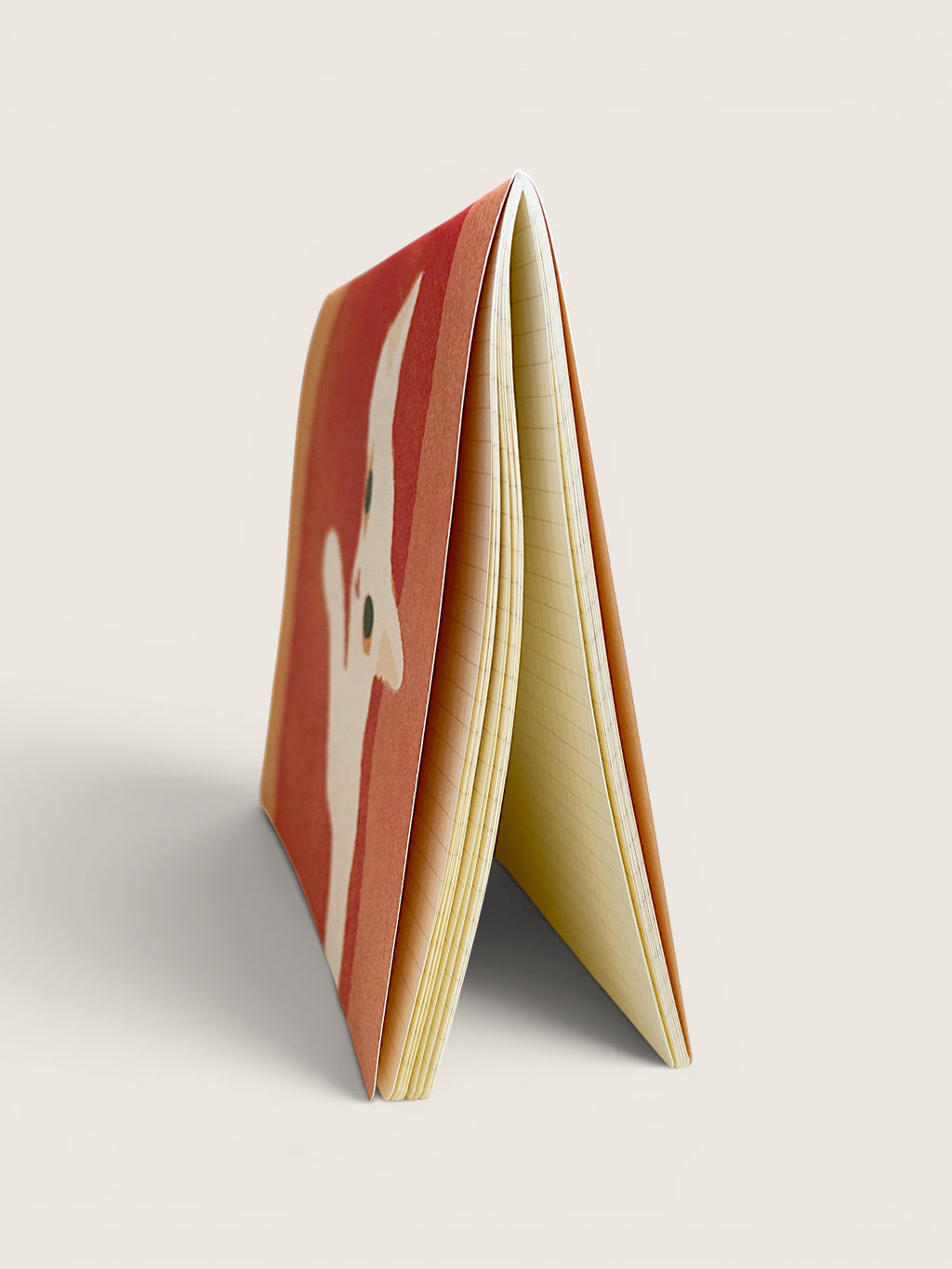 A thin, flexible notebook or card stack standing upright at an angle, showing a red-orange cover with a white stylized face illustration and cream-colored pages on a light beige background.