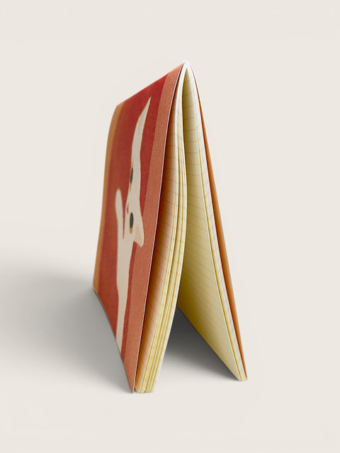 A thin, flexible notebook or card stack standing upright at an angle, showing a red-orange cover with a white stylized face illustration and cream-colored pages on a light beige background.