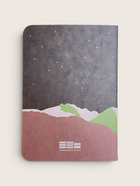 A Yamahito Paper notebook with a minimalist landscape cover design, featuring a dark starry night sky, green and white mountains, and a dusty rose foreground, shown on a light beige background.