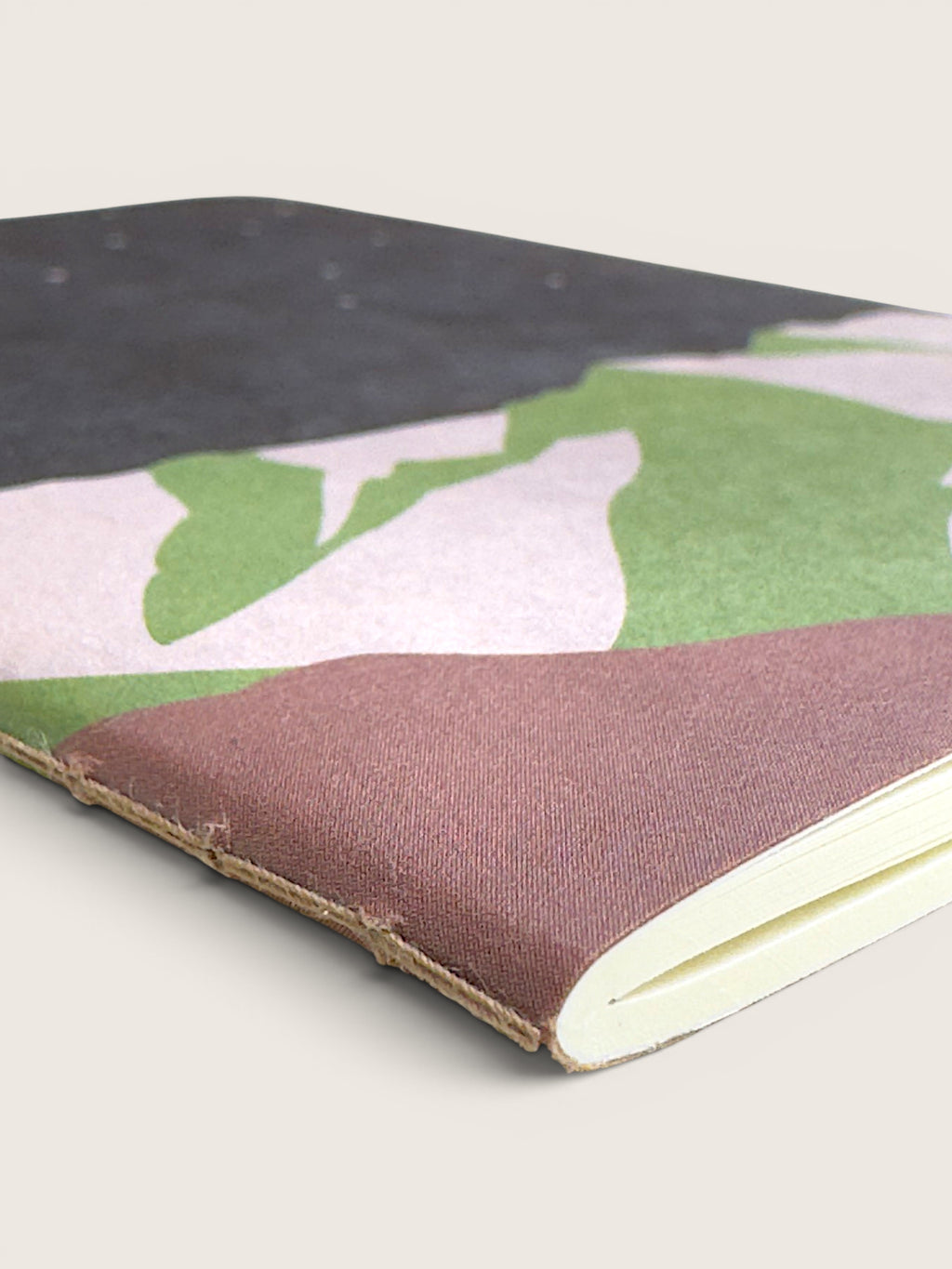 A close-up of a notebook with a camouflage-patterned cover in green, pink, and black, featuring a rustic brown leather binding and cream-colored pages.