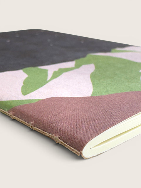A close-up of a notebook with a camouflage-patterned cover in green, pink, and black, featuring a rustic brown leather binding and cream-colored pages.