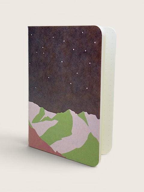 A hardcover notebook with a minimalist mountain and starry night design, standing upright on a light beige background.