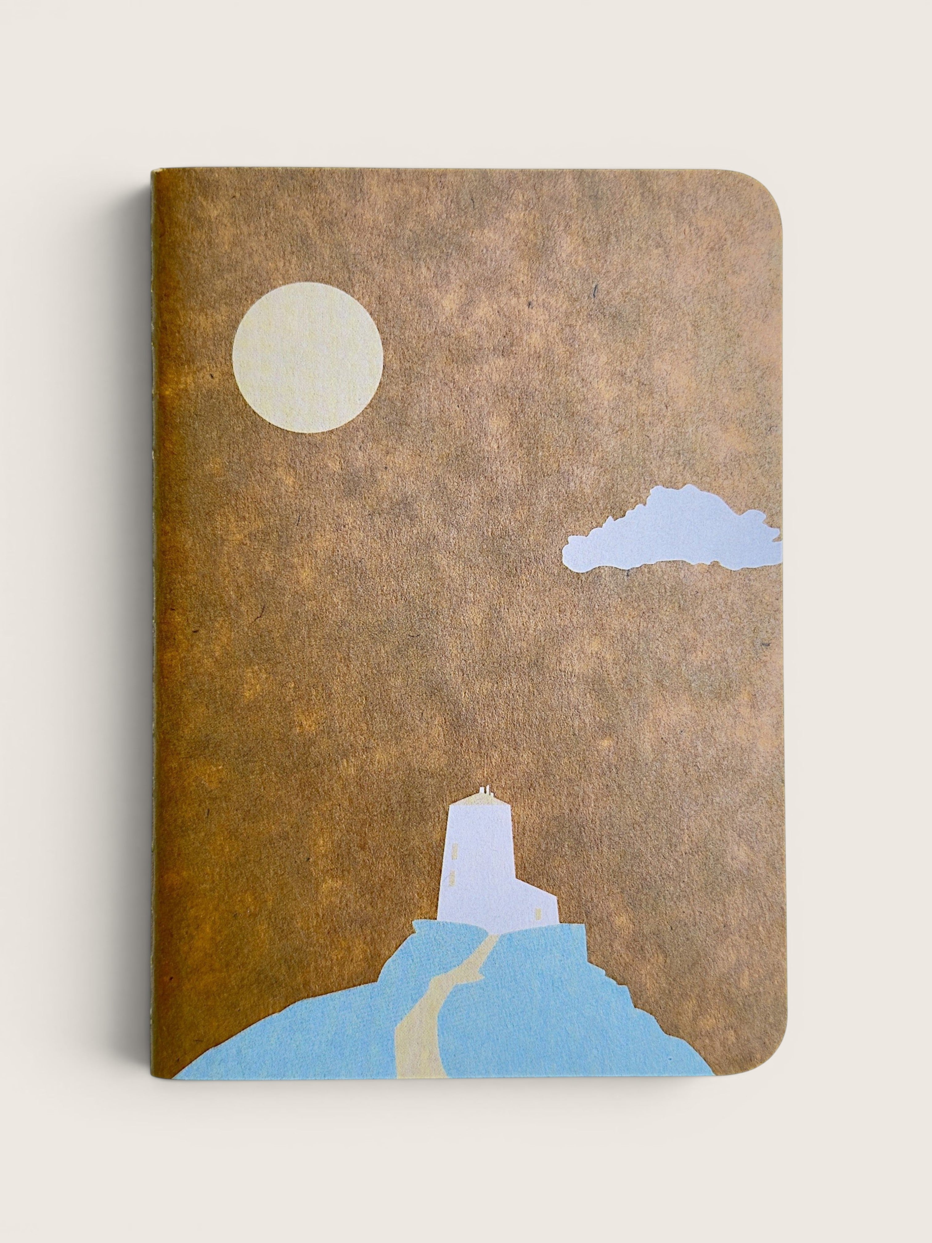 A notebook with a textured brown cover featuring a minimalist illustration of a lighthouse on a blue hill under a cream moon and light blue cloud, shown on a light beige background.