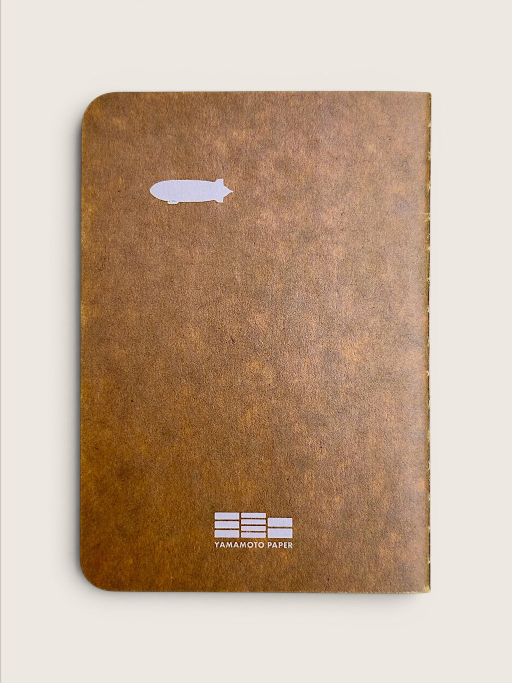 A brown textured notebook with a small white blimp graphic near the top and a white Yamamoto Paper logo at the bottom, shown on a light beige background.