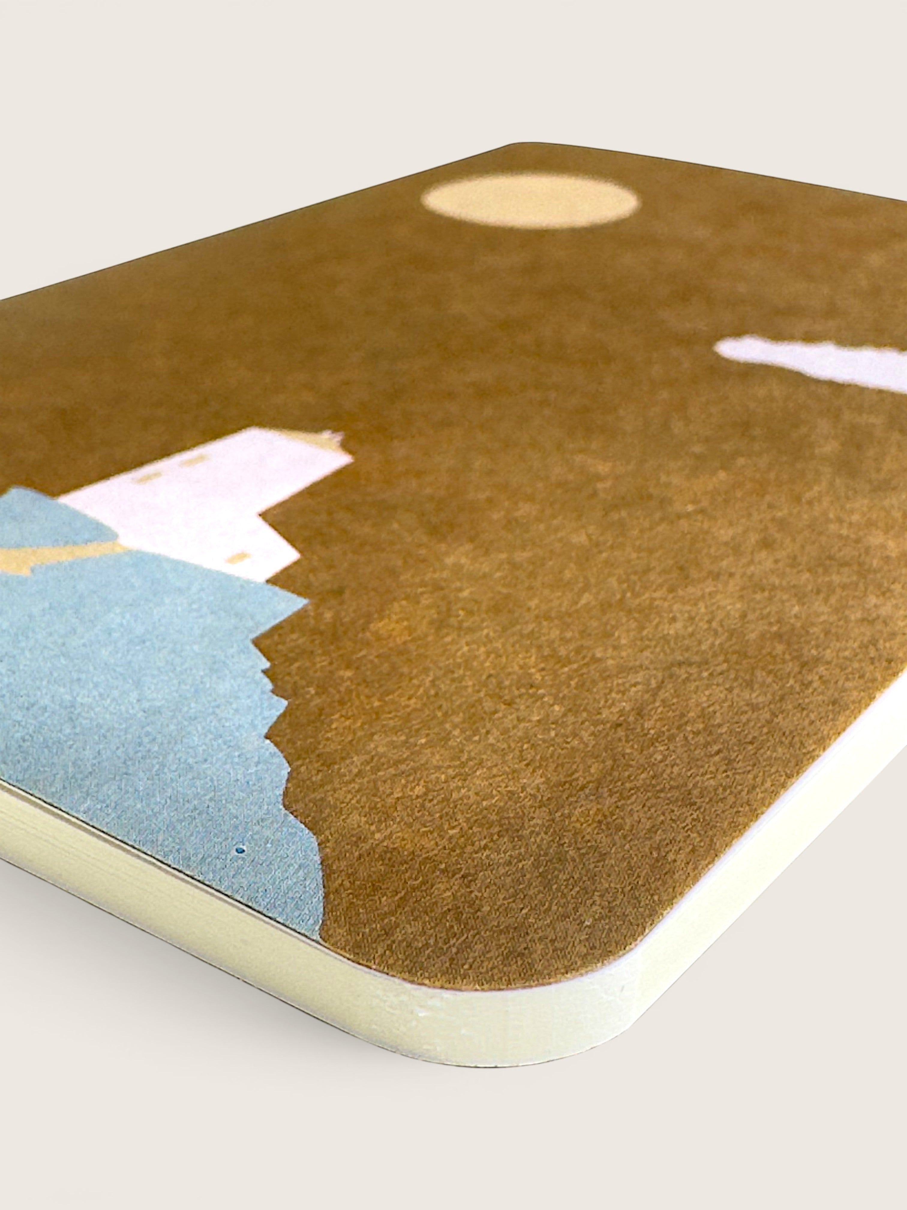 A close-up of a rectangular desk mat or mousepad with rounded corners, featuring a textured brown background with abstract paper cutouts in light blue, pale pink, cream, and gold. The product is shown at an angle on a light beige background.