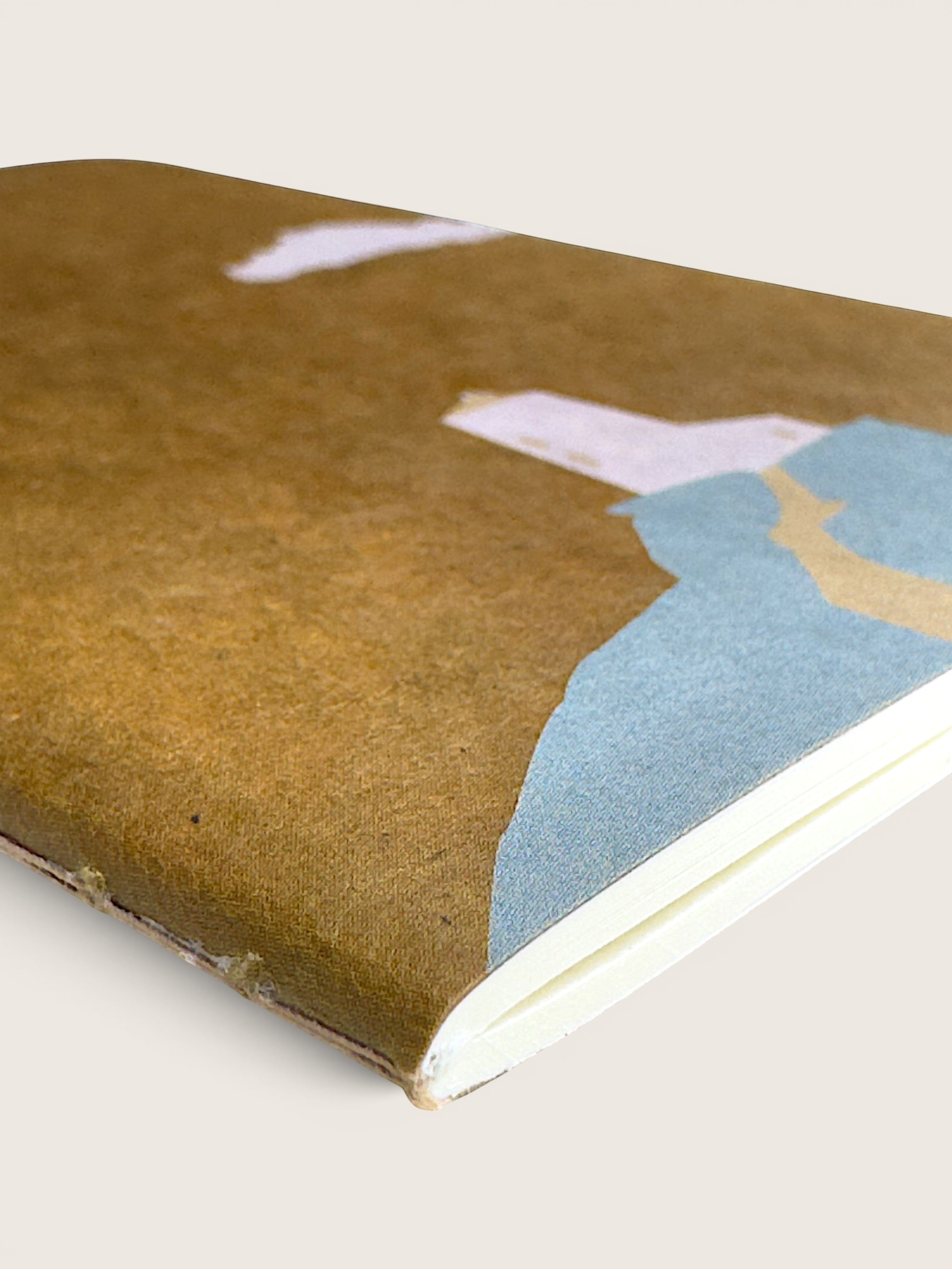 A close-up of a notebook with a textured brown cover featuring a minimalist abstract landscape design in light blue, beige, and white, set against a solid light beige background.