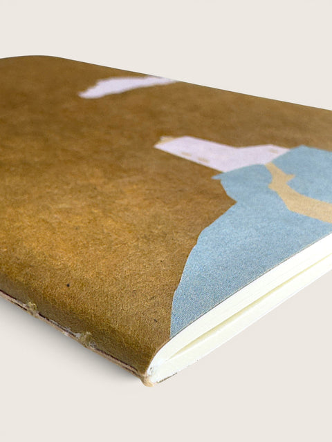 A close-up of a notebook with a textured brown cover featuring a minimalist abstract landscape design in light blue, beige, and white, set against a solid light beige background.